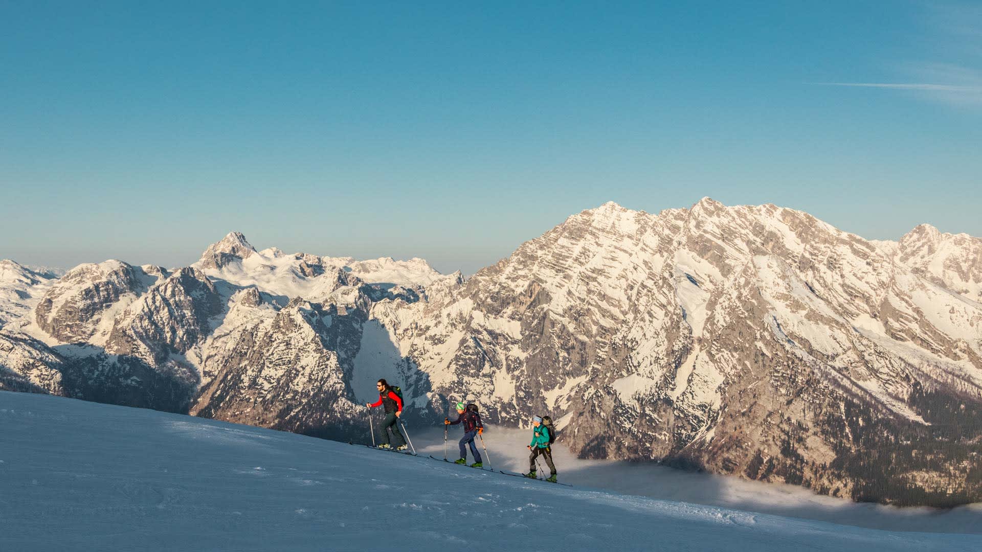 Drei Skitourengeher steigen mit Fellen auf einem schneebedeckten Hang in den Berchtesgadener Alpen auf.