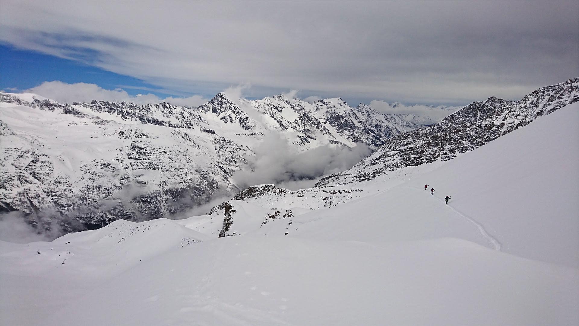 Drei Skitourengeher im Aufstieg auf flachem Schneehang, im Hintergrund die Gran-Paradiso-Bergkette mit wolkenverhüllten Gipfeln.