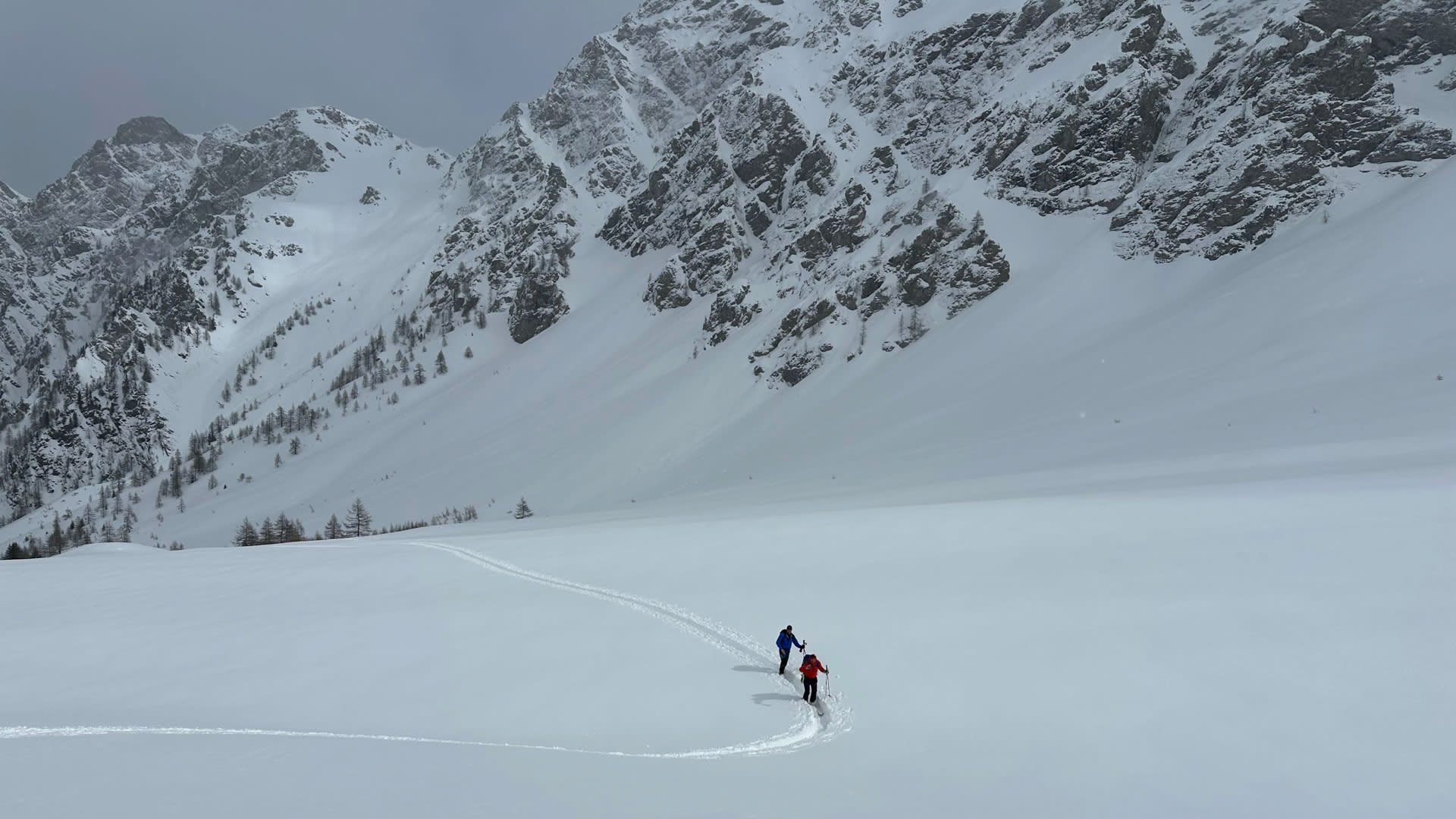Zwei Skitourengeher hinterlassen geschwungene Spuren im Tiefschnee am Großen St. Bernhard vor einer Felswand.