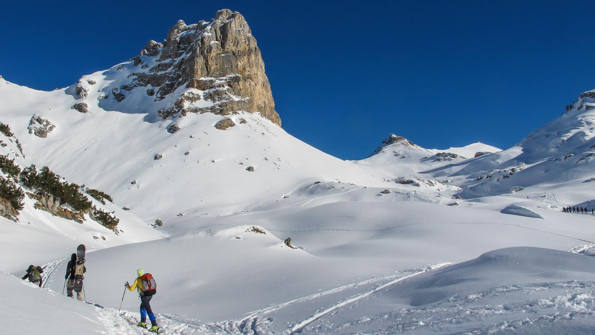 Skitourengeher steigen im Rofangebirge einen verschneiten Hang auf, im Hintergrund ein markanter Kalksteinfelsturm.
