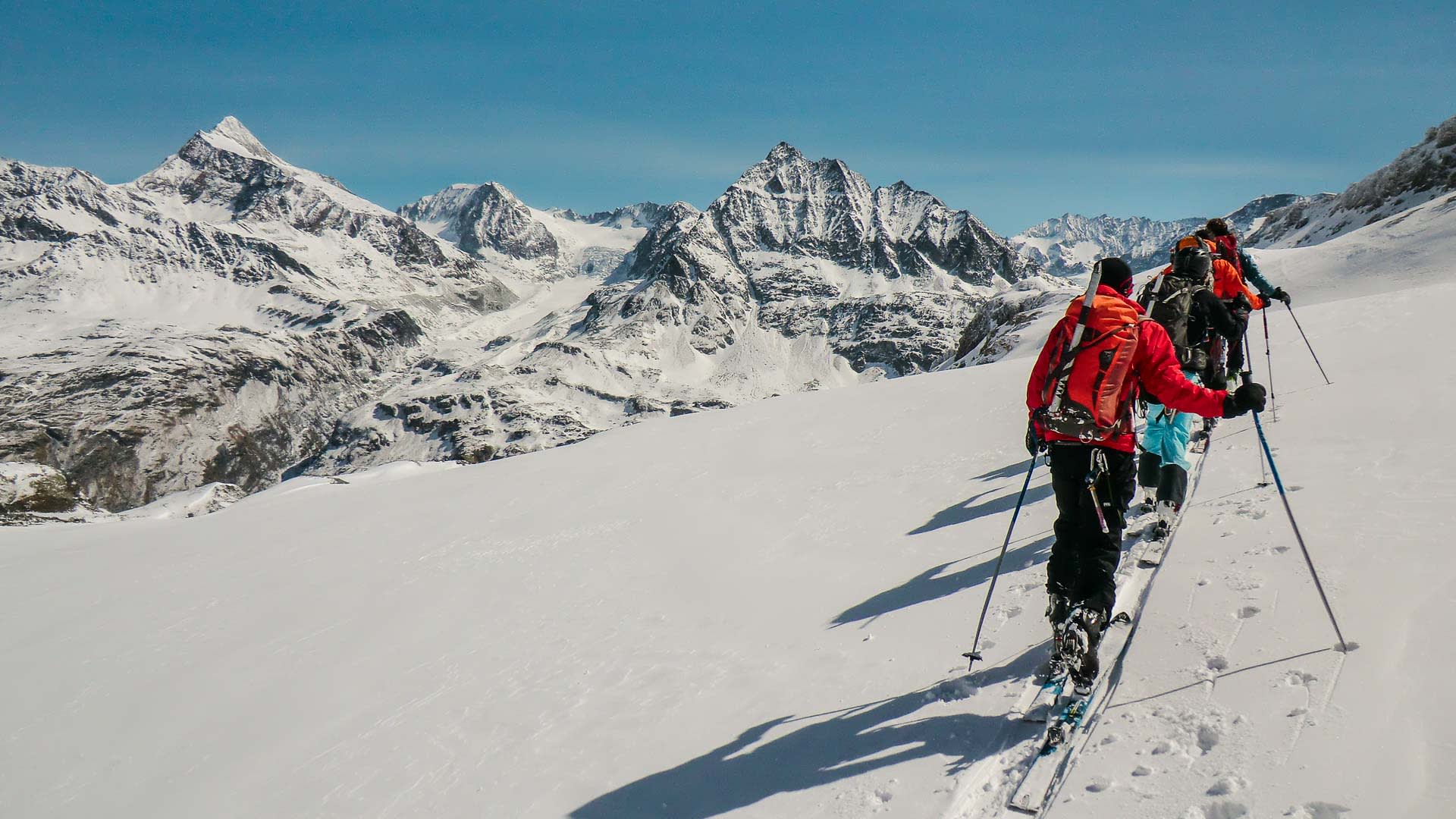 Drei Skitourengeher mit Fellen steigen in einer Linie einen verschneiten Hang im Wallis auf.