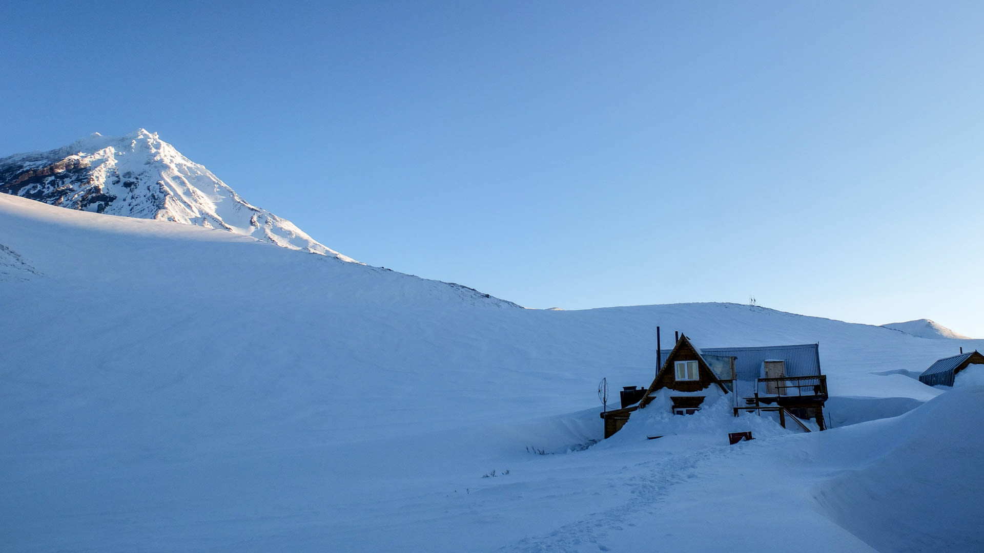Holzgebäude des Avacha Camps unter Schneelast mit dem spitzen Avacha-Gipfel im Hintergrund bei blauem Winterhimmel.