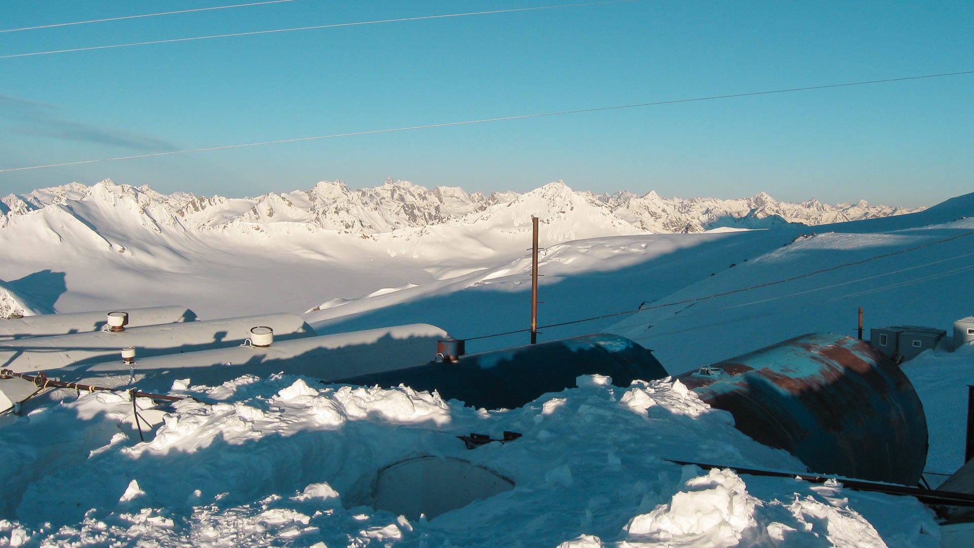 Skitour-Basislager am Elbrus: verschneites Hochplateau mit technischen Anlagen und vergletscherter Bergkette im Hintergrund.