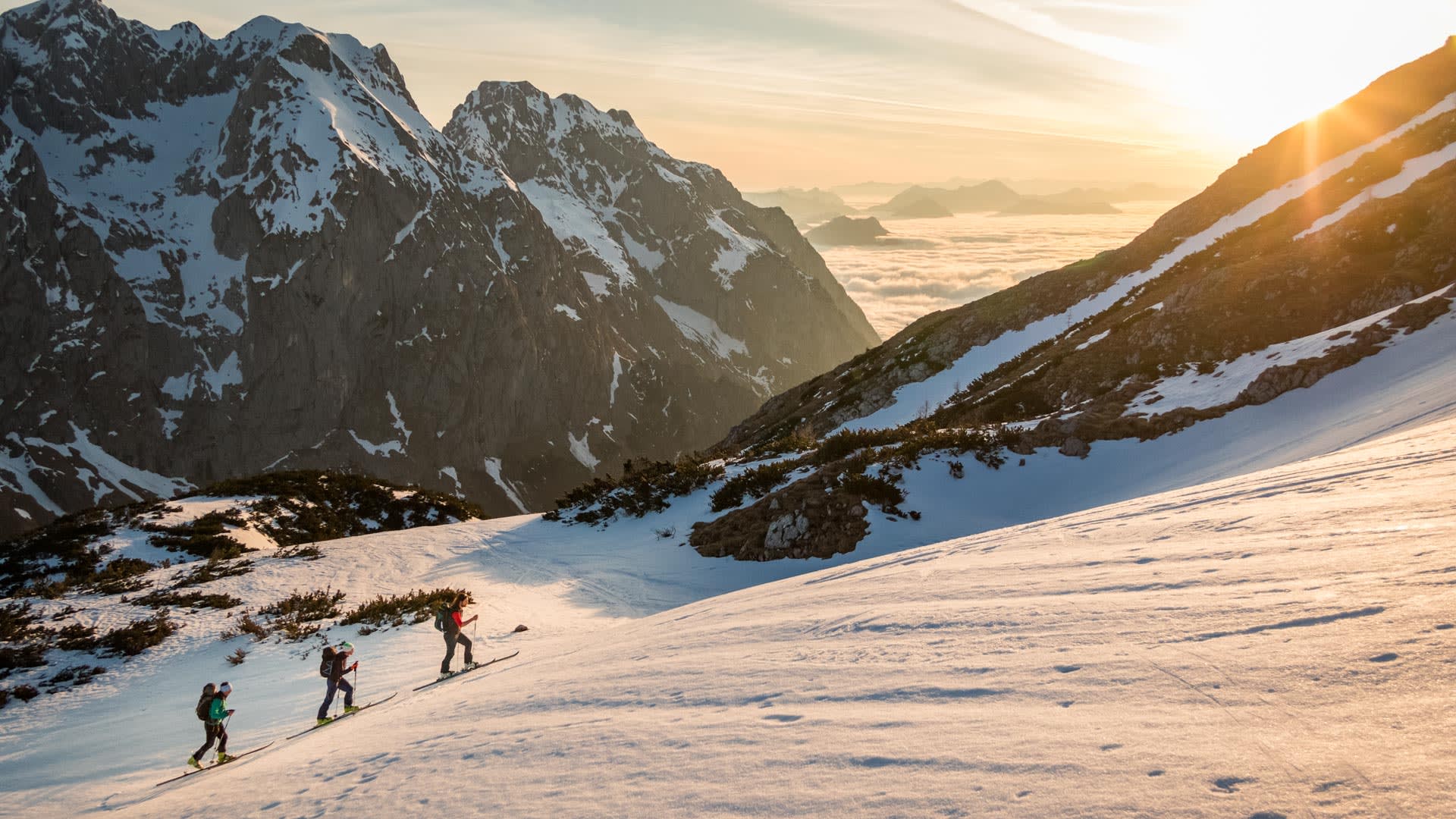 Drei Skitourengeher steigen diagonal einen Schneehang in den Berchtesgadener Alpen auf, im Hintergrund steile Felsgipfel