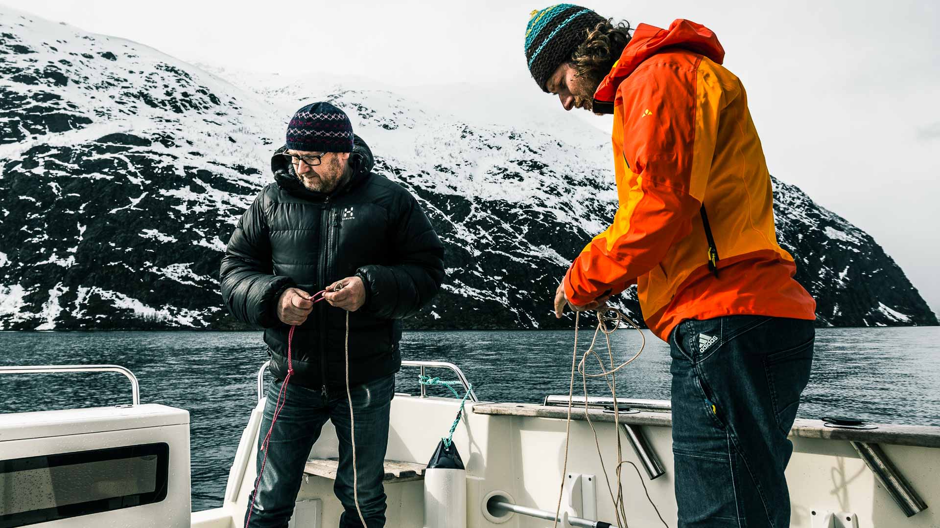 Zwei Skitourengeher hantieren mit Seilen auf dem Heck eines Motorboots vor einem verschneiten Fjordberg in Norwegen.