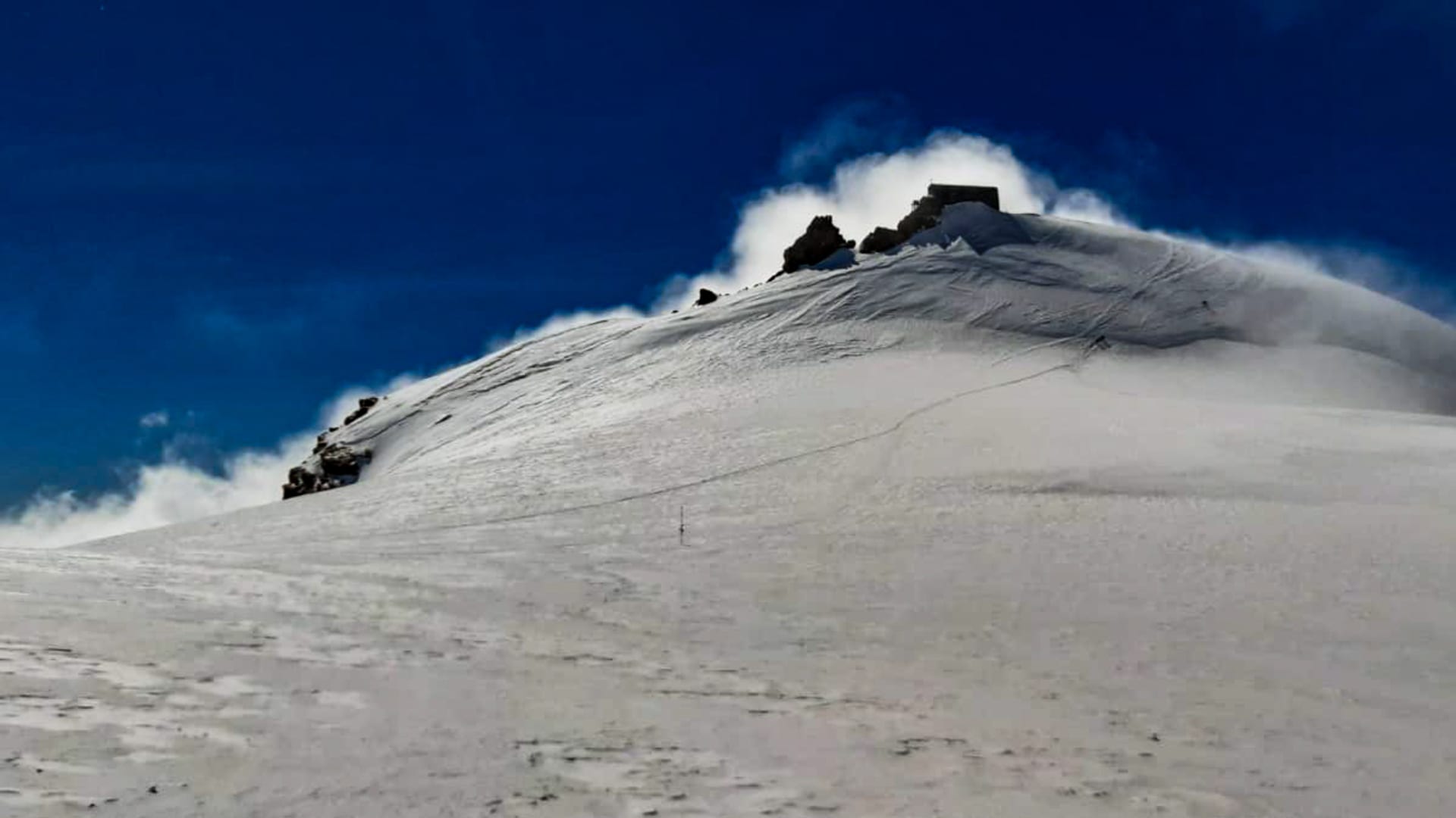 Skitour zur Capanna Margherita: schneebedeckter Steilhang mit Berghütte am Gipfel und dunkelblauen Himmel.