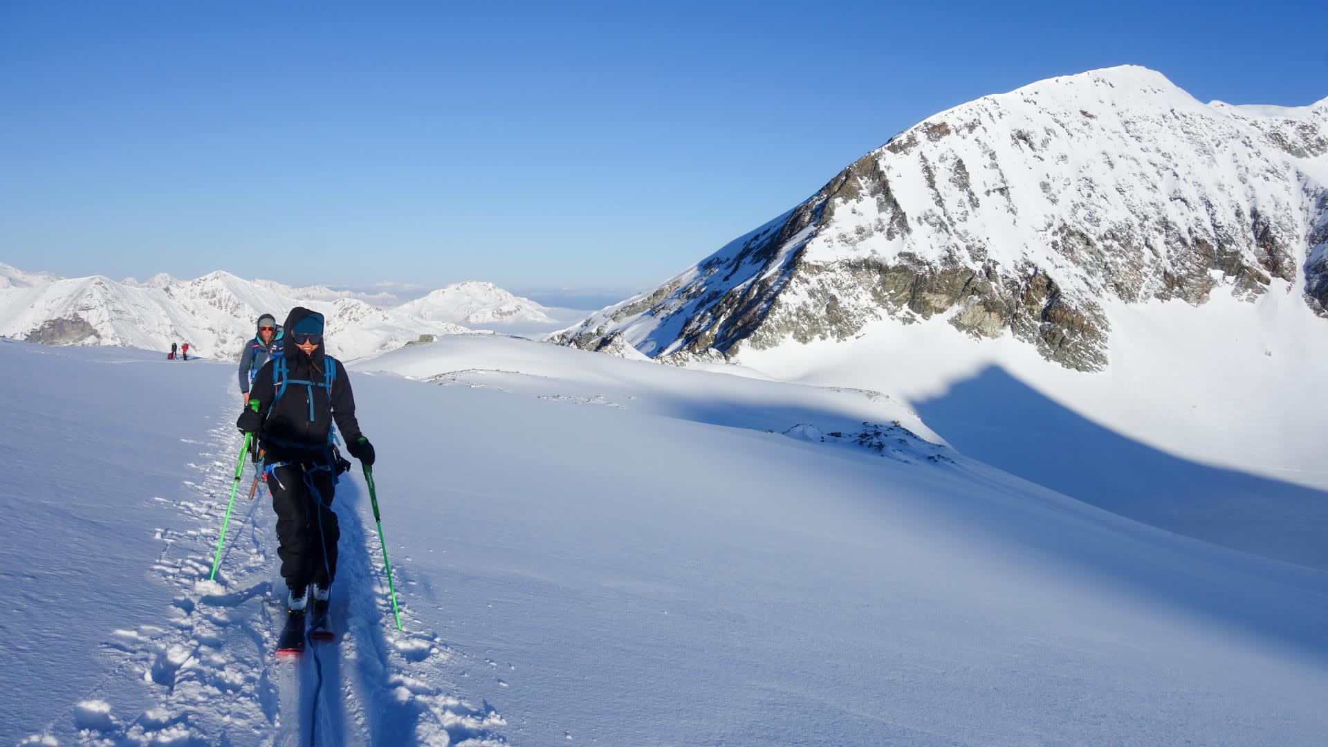 Zwei Skitourengeher mit Fellen steigen einen breiten Schneehang im Wallis auf, rechts ein verschneiter Gipfel.