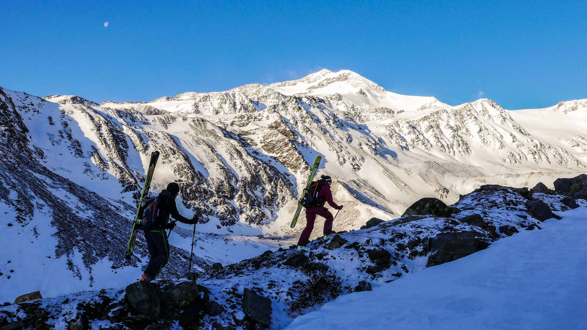 Zwei Skitourengeher traversieren einen felsig-schneeigen Grat am Cevedale bei tiefblauem Himmel mit Mondschel.
