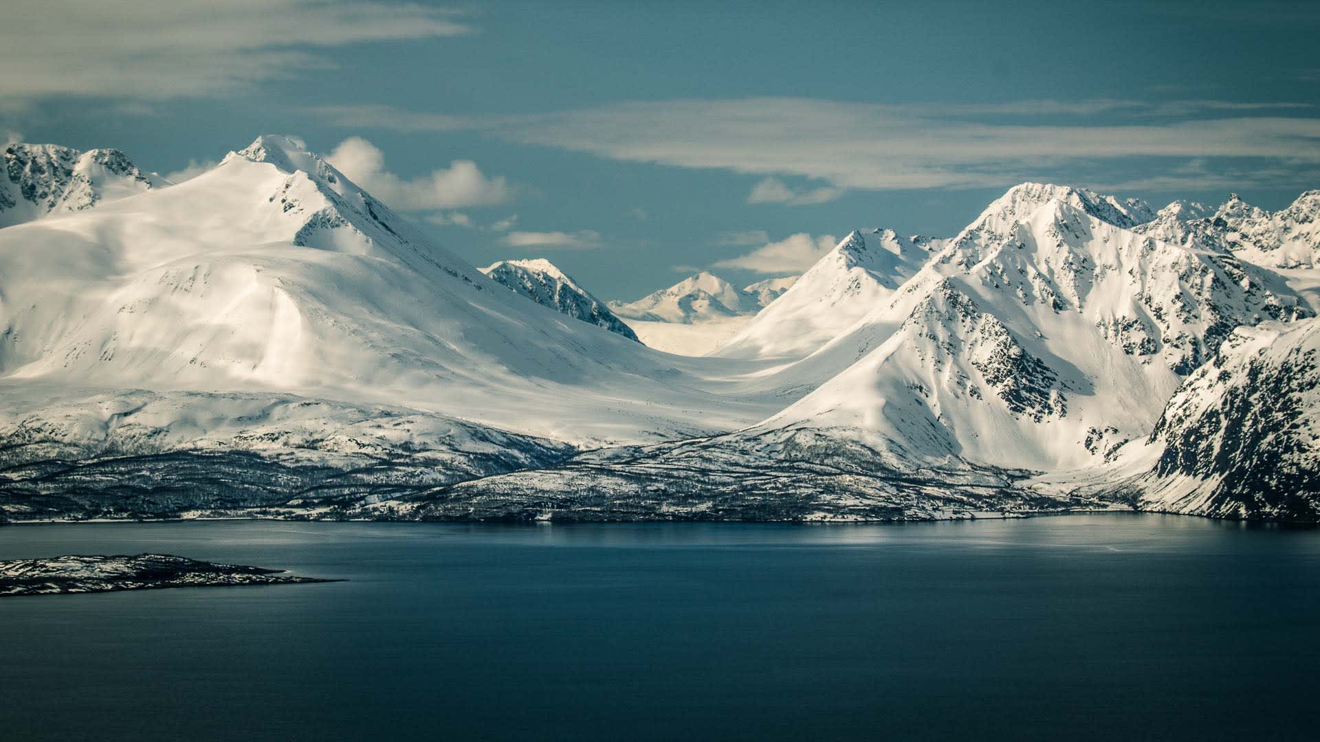 Winterliche Fjordlandschaft in Norwegen mit schneebedeckten Gipfeln bis auf Meereshöhe und ruhiger, tiefblauer Wasseroberfläche.