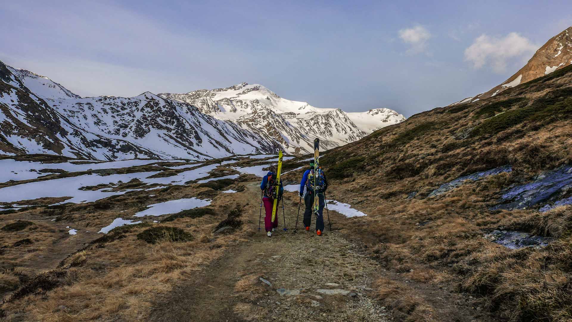 Zwei Skitourengeher steigen mit Ski auf dem Rücken einen Wanderweg ins Tal Richtung Cevedale auf.