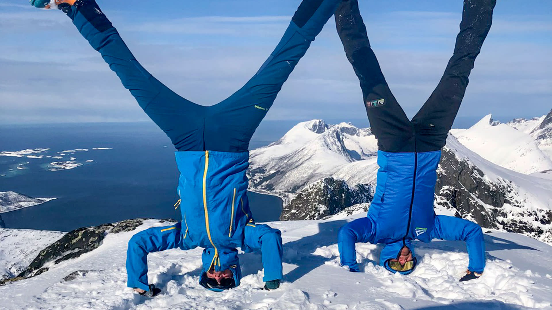Zwei Skitourengeher machen einen Kopfstand auf dem verschneiten Gipfel in Senja Norwegen, Fjord und Bergkämme im Hintergrund.