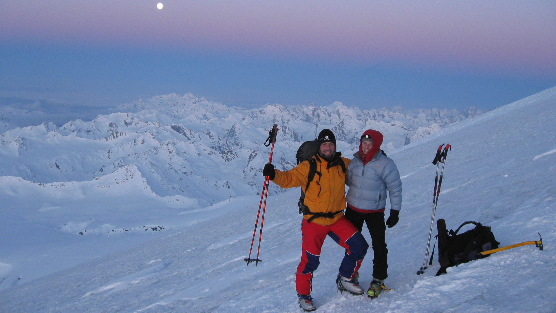 Zwei Skitourengeher mit Steigeisen und Skistöcken am Steilhang des Elbrus bei Dämmerung, Vollmond und Hochgebirgspanorama