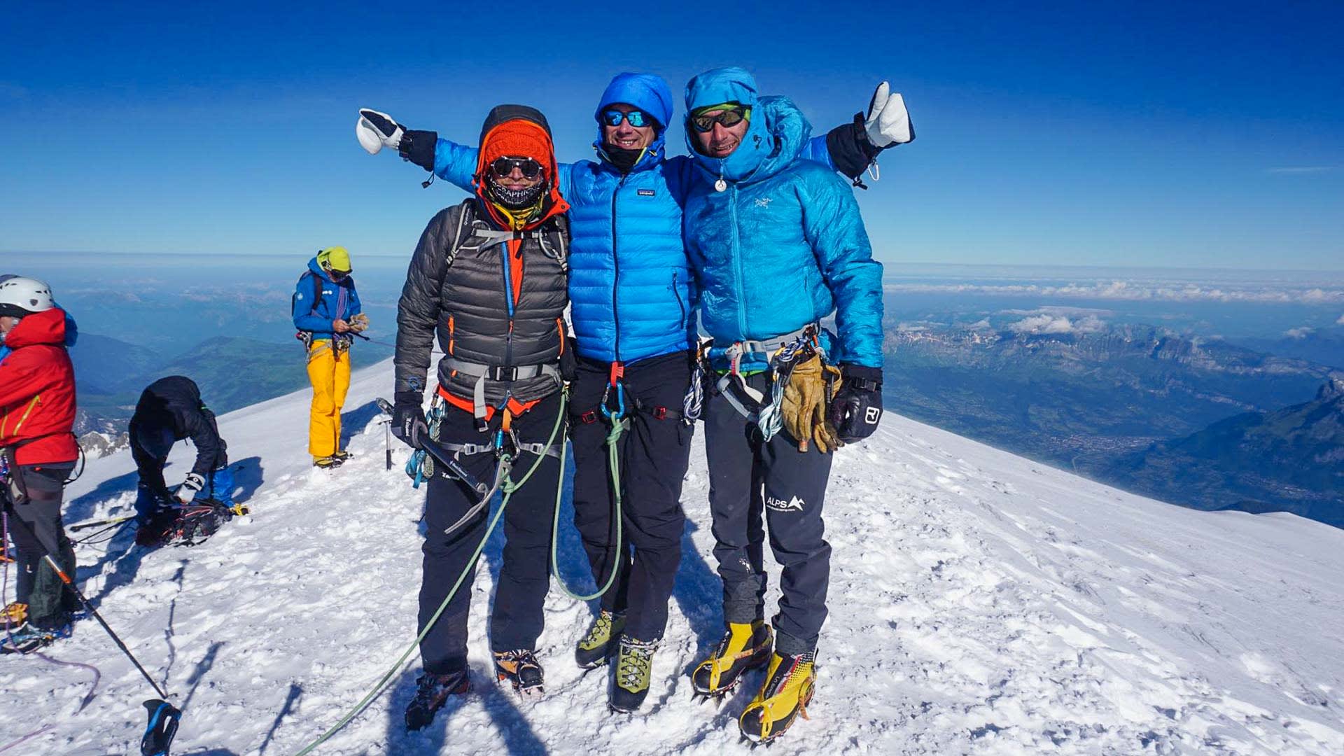Drei Bergsteiger mit Steigeisen und Klettergurten posieren auf einem schneebedeckten Gipfelgrat im Mont-Blanc-Massiv.