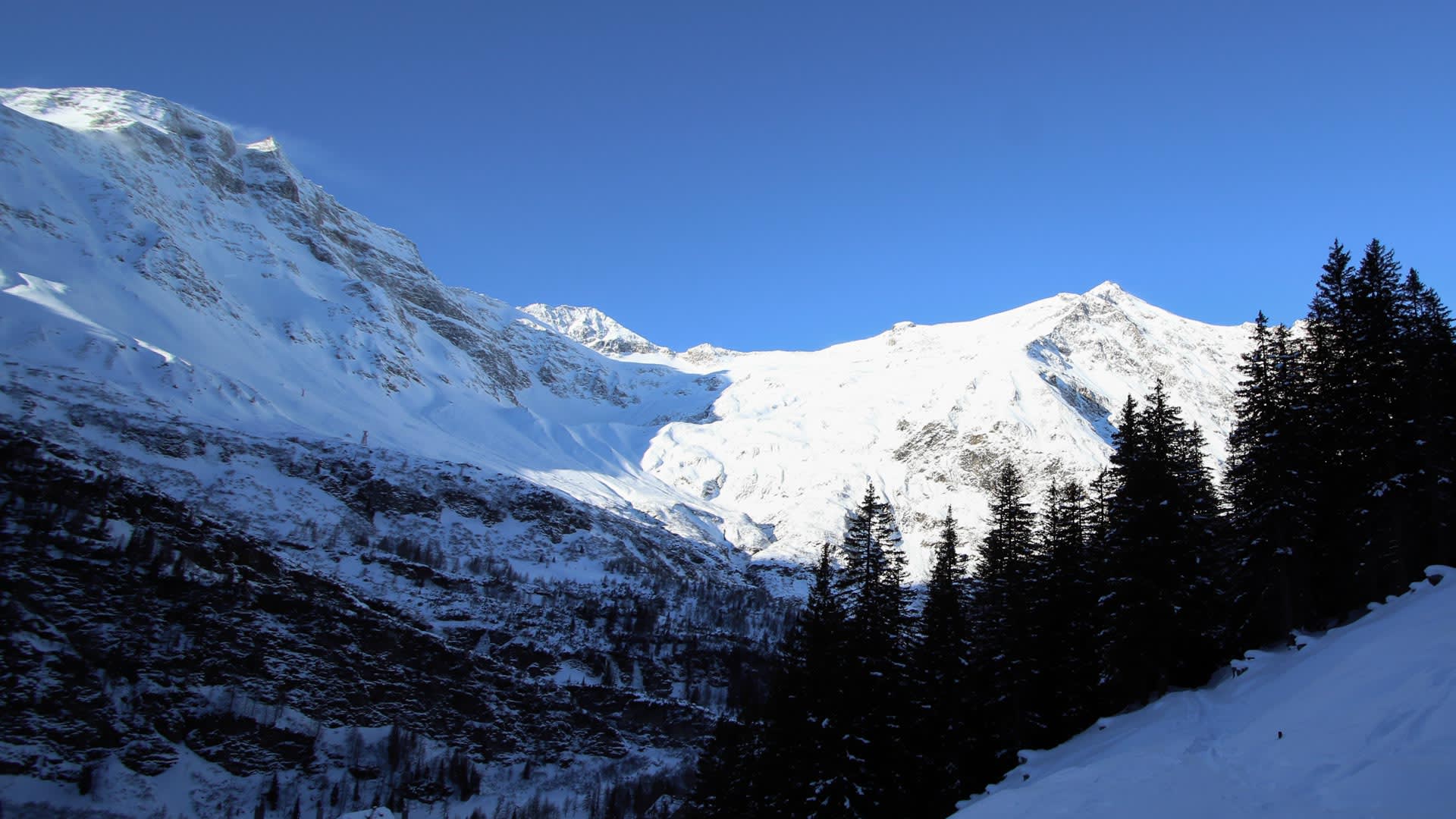 Skitour an der Sonnblickbasis: zwei verschneite Gipfel über einem Talkessel mit Nadelbäumen im Vordergrund.