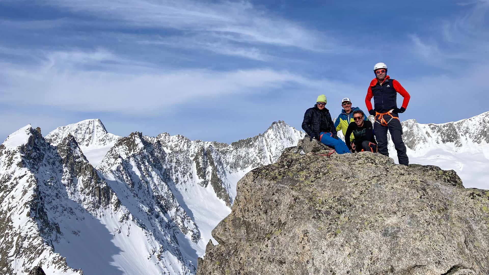 Vier Skitourengeher rasten auf einem felsigen Gipfelblock mit weitem Hochgebirgspanorama der Urner Haute Route.