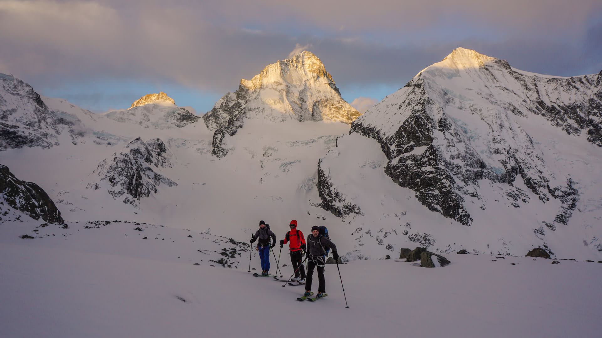 Drei Skitourengeher mit Fellen und Rucksäcken stehen auf einem Gletscherplateau im Wallis vor einem pyramidenförmigen Gipfel.