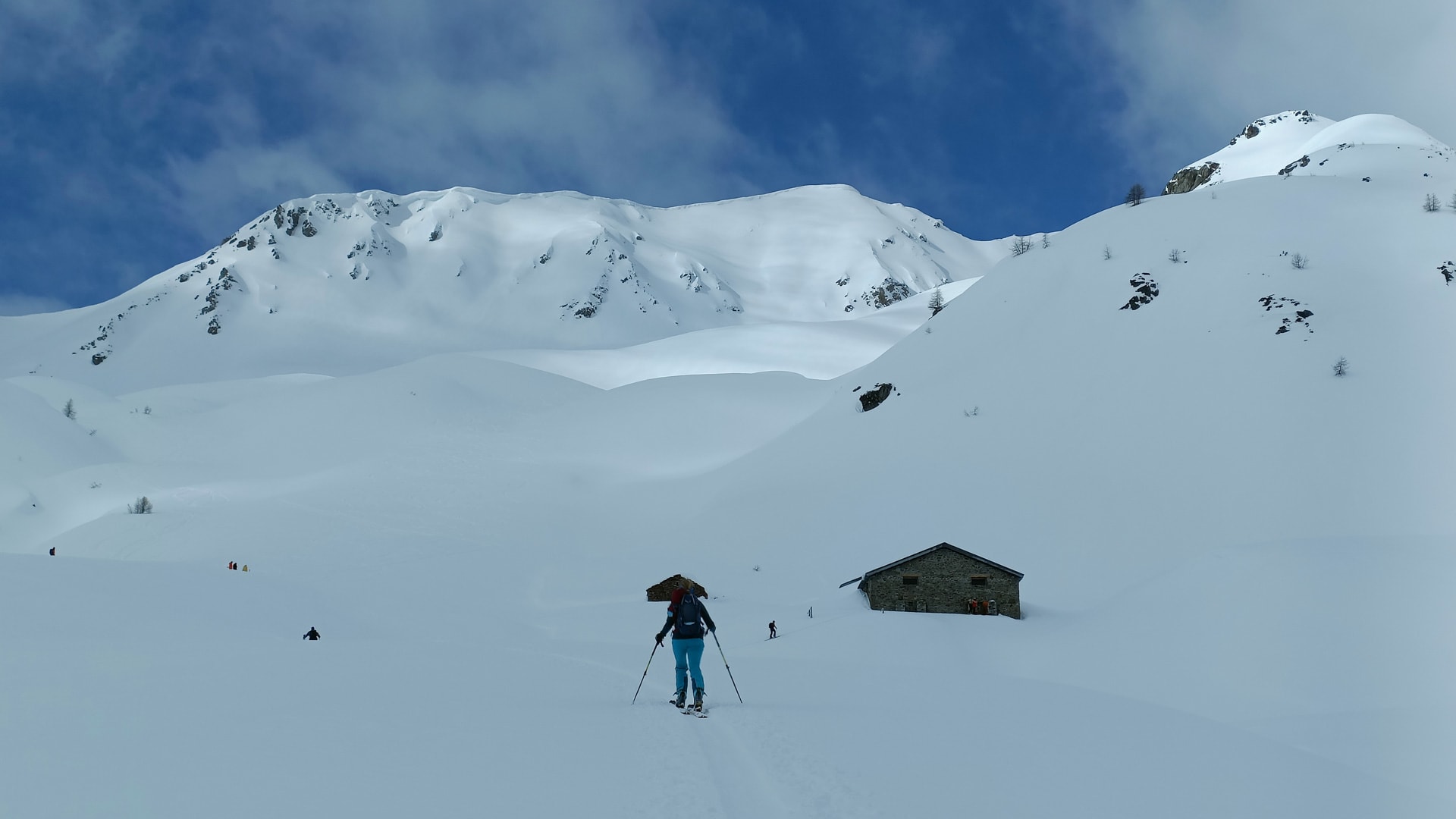 Skitourengeher mit Fellen und Rucksack steigt am Großen St. Bernhard an einer Steinhütte vorbei auf.