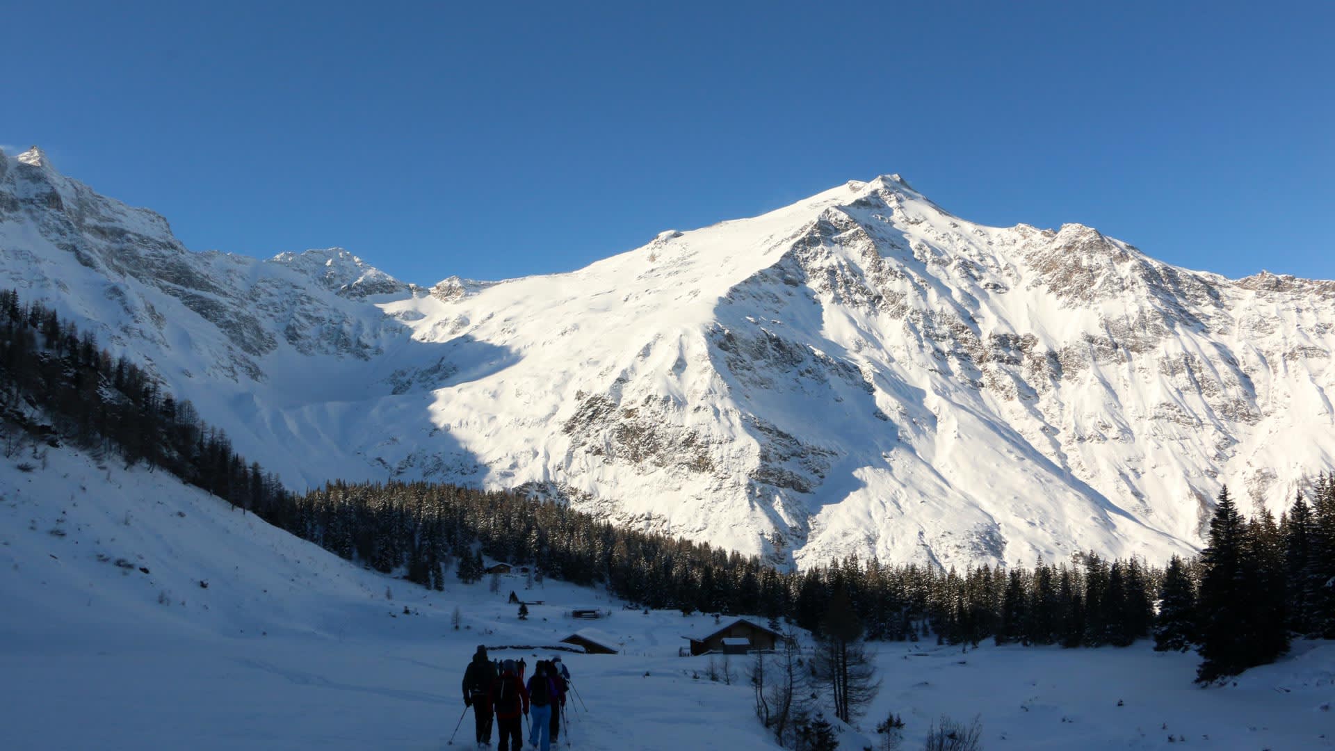 Fünf Skitourengeher stehen im verschneiten Rauriser Tal vor Almhütten und einem massiven Gipfel im Hintergrund.
