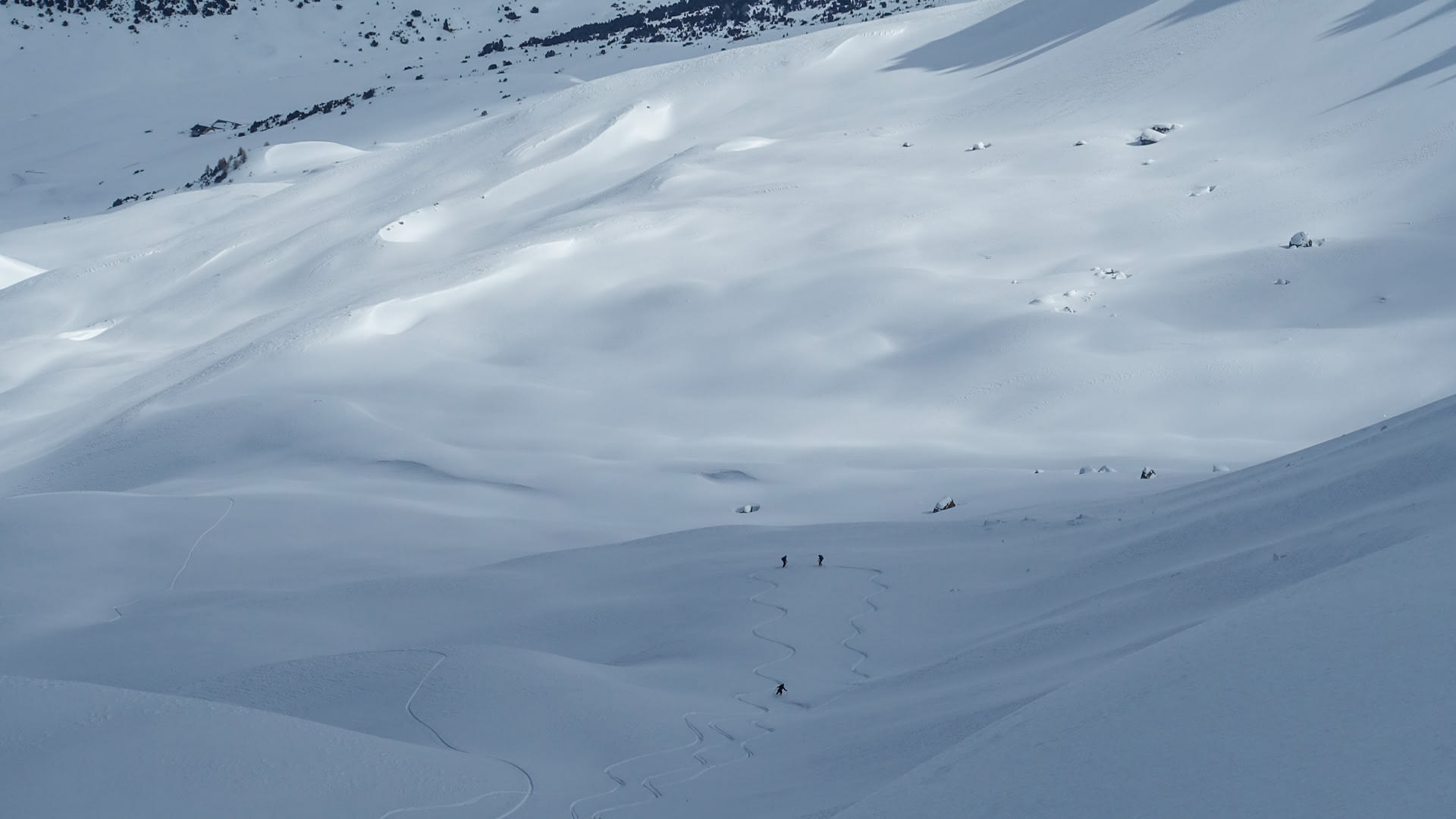 Drei Skitourengeher auf einem ausgedehnten Pulverschneehang im Val Müstair, windgeformte Schneestrukturen und Felsbrocken sichtbar.
