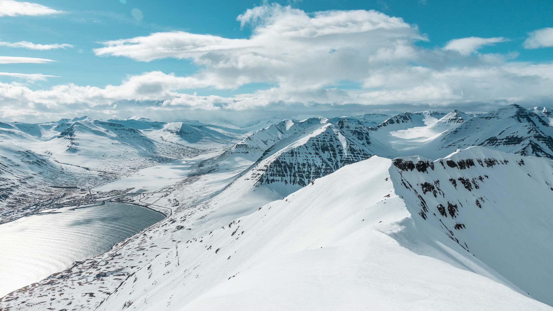 Skitour auf Island: Panoramablick vom Berggrat auf verschneite Gipfel, Fjord und eine kleine Talsiedlung unter Cumuluswolken.