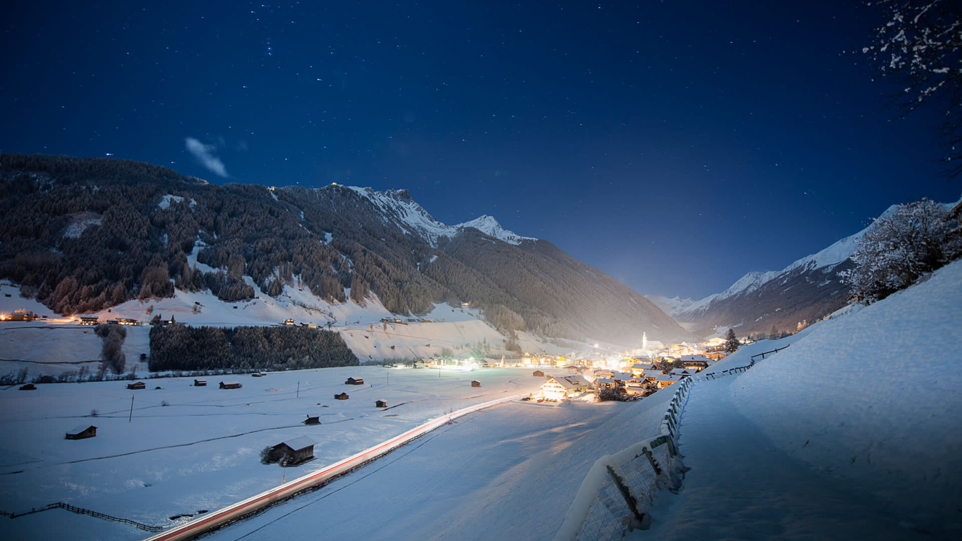 Langzeitbelichtung von Neustift im Stubaital bei Nacht mit Lichtschlieren auf der Talstraße und beleuchtetem Kirchturm.