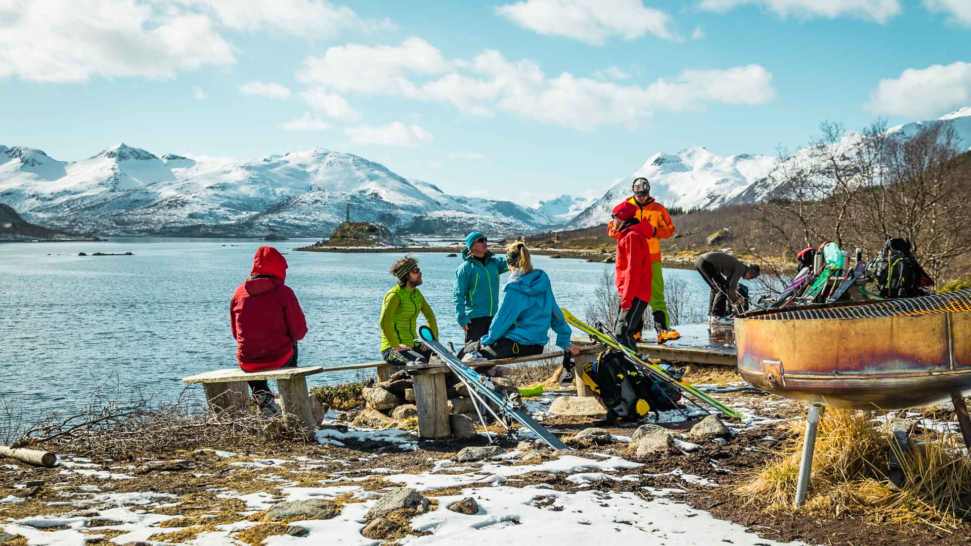 Skitourengehergruppe pausiert auf einer Bank am Fjordufer der Lofoten mit Ski und Bergen im Hintergrund.