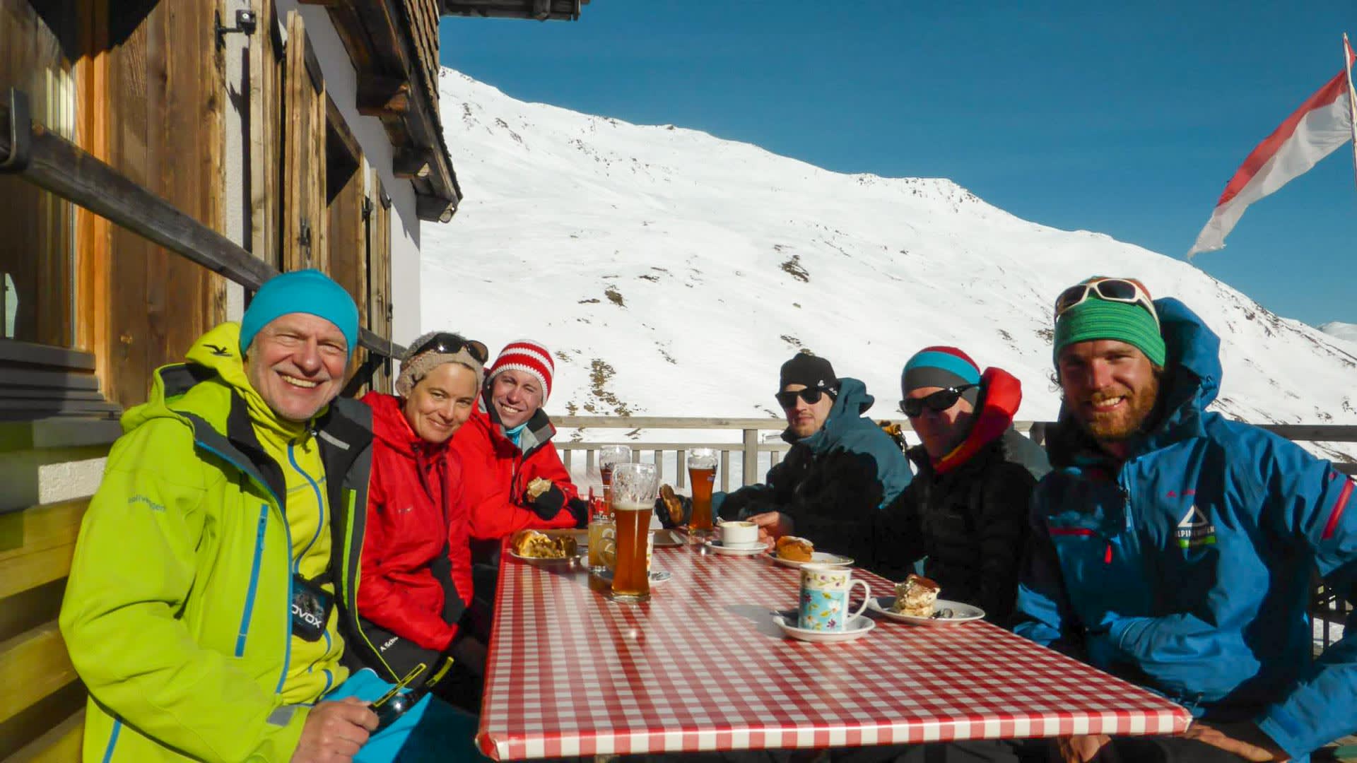 Sechs Skitourengeher genießen eine Rast auf der sonnigen Außenterrasse der Sesvennahütte mit Bier, Kaffee und Kuchen