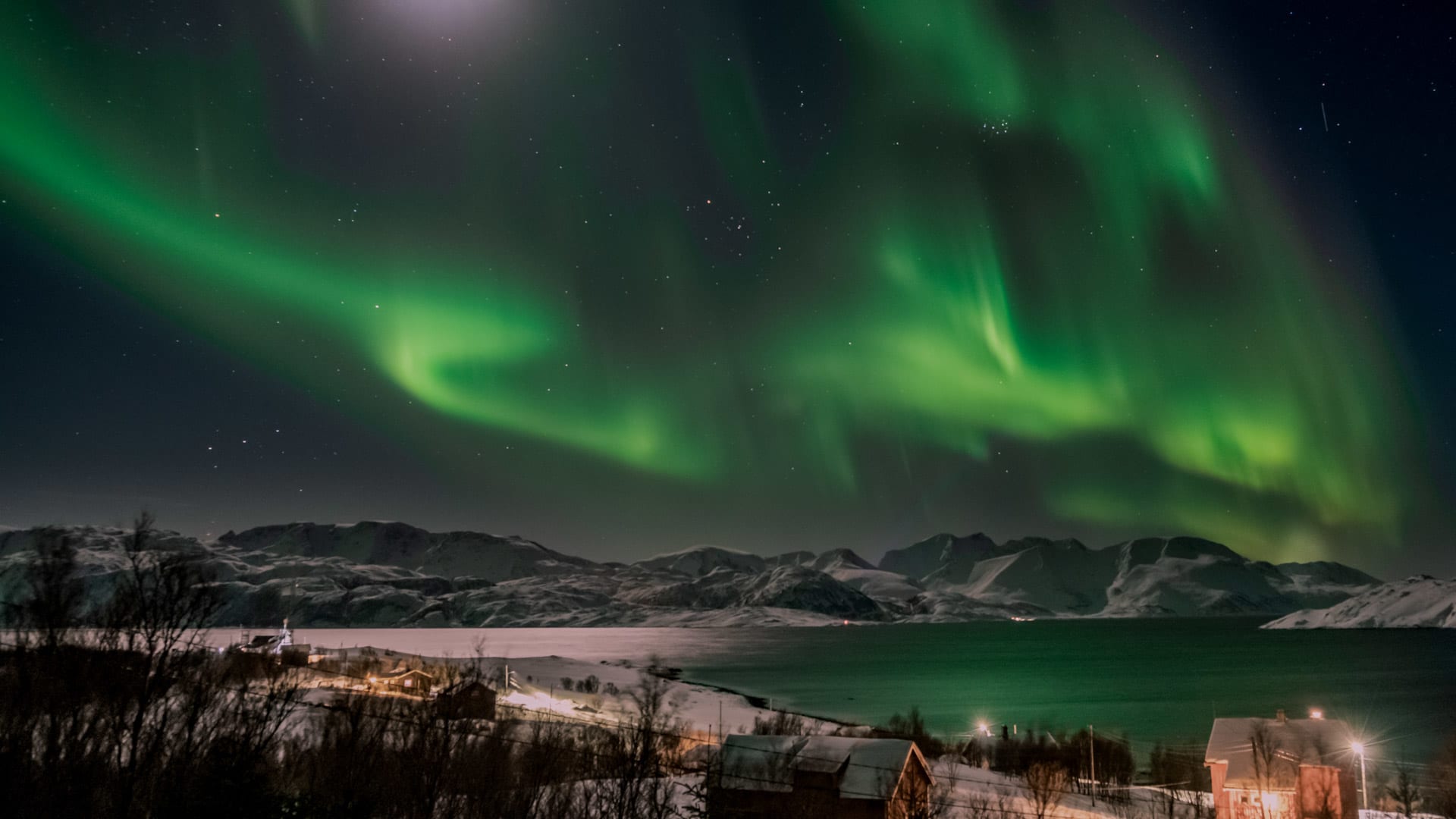 Skitour Seiland: Nordlichter in grünen Bögen über schneebedeckten Bergen und beleuchtetem Dorf am Fjord.