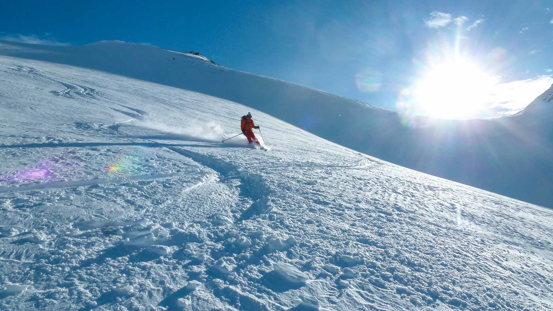 Skitourengeher in oranger Jacke fährt mit Pulverschneewolke einen Hang bei der Tuoi Hütte in der Silvretta ab.