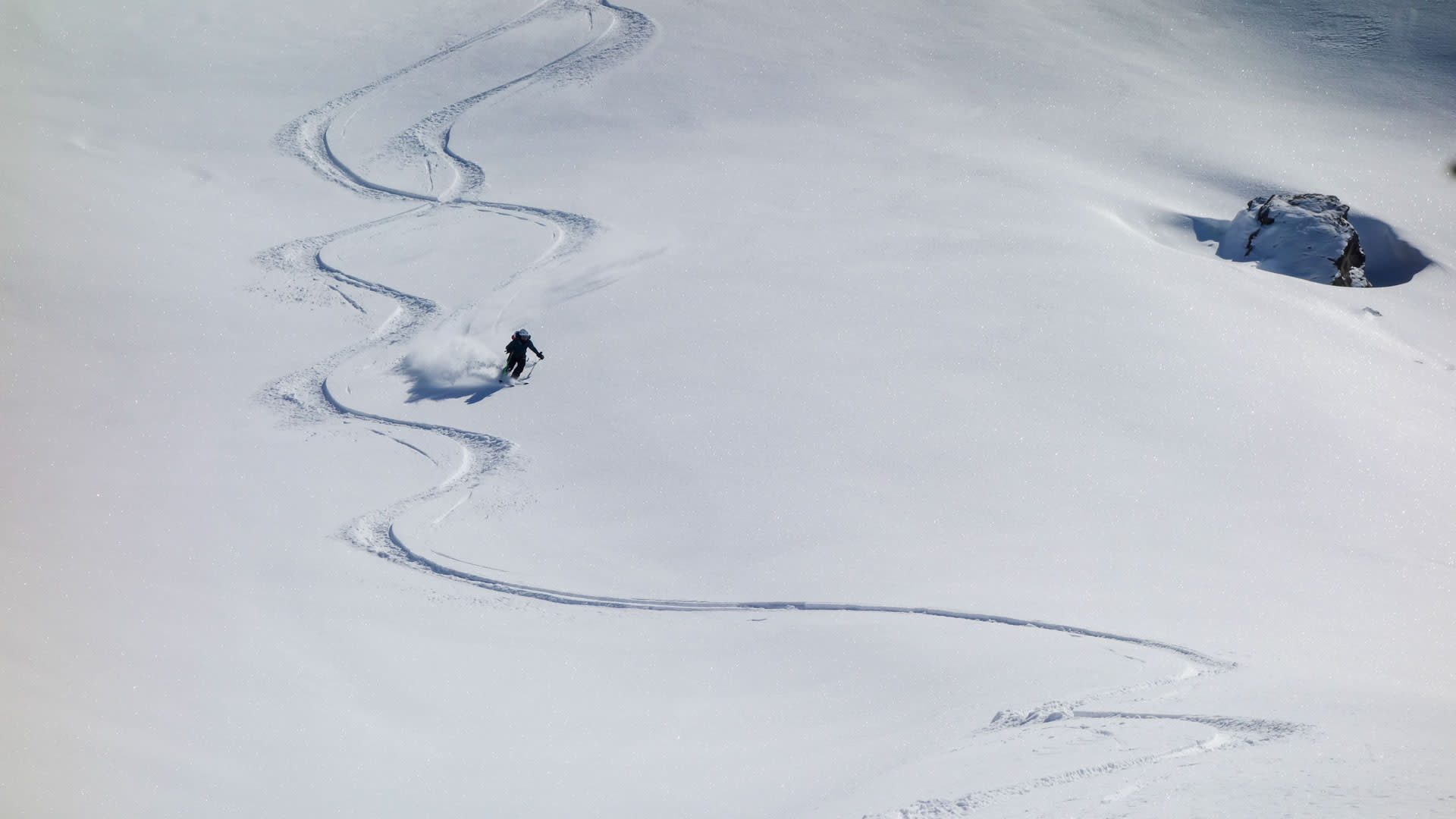 Ein Skitourengeher fährt einen unberührten Pulverschneehang im Wallis ab, mit geschwungenen Spuren und aufgewirbelter Schneewolke.