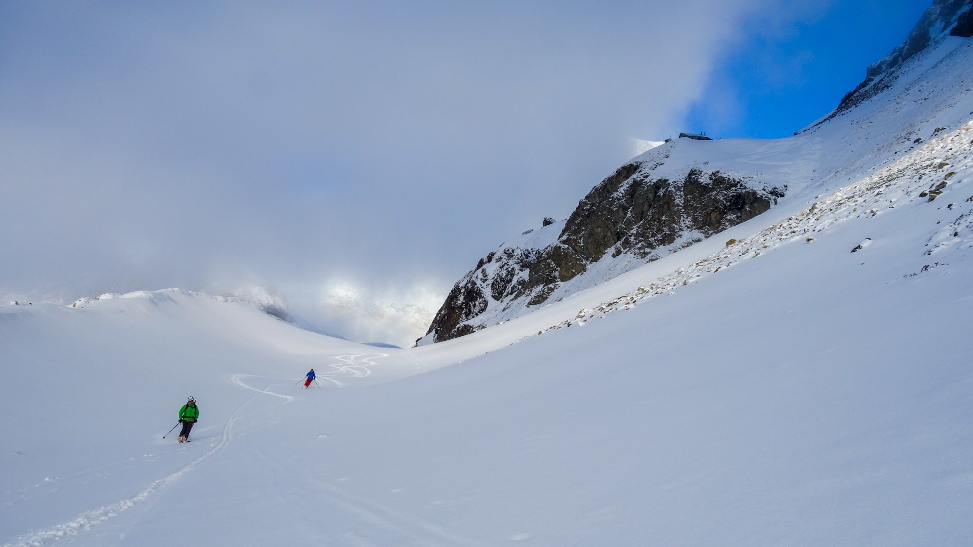 Zwei Skitourengeher fahren auf einem weitläufigen Pulverschneehang im Wallis ab, rechts ein dunkler Felsberg mit Berghütte.