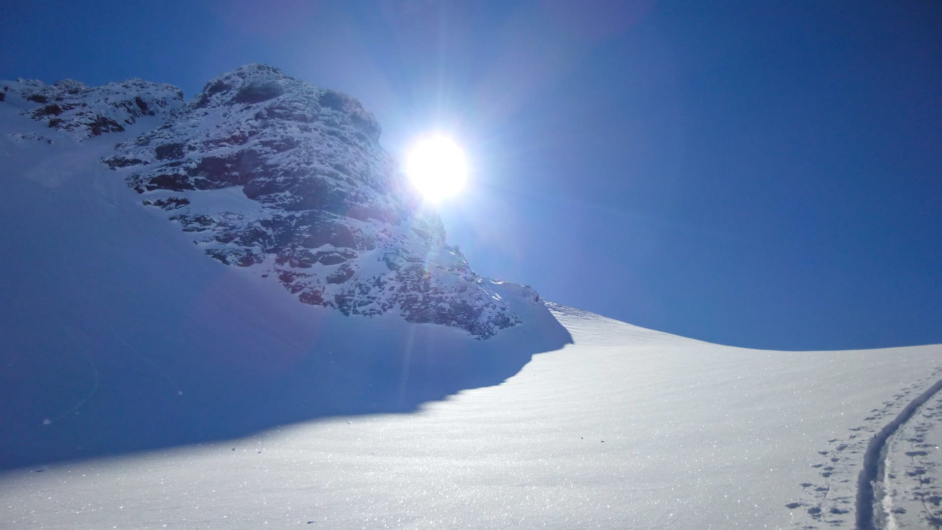 Skitour zur Rauhen-Kopf-Scharte: Skispuren im Gegenlicht auf weitem Schneehang mit felsiger Bergflanke.