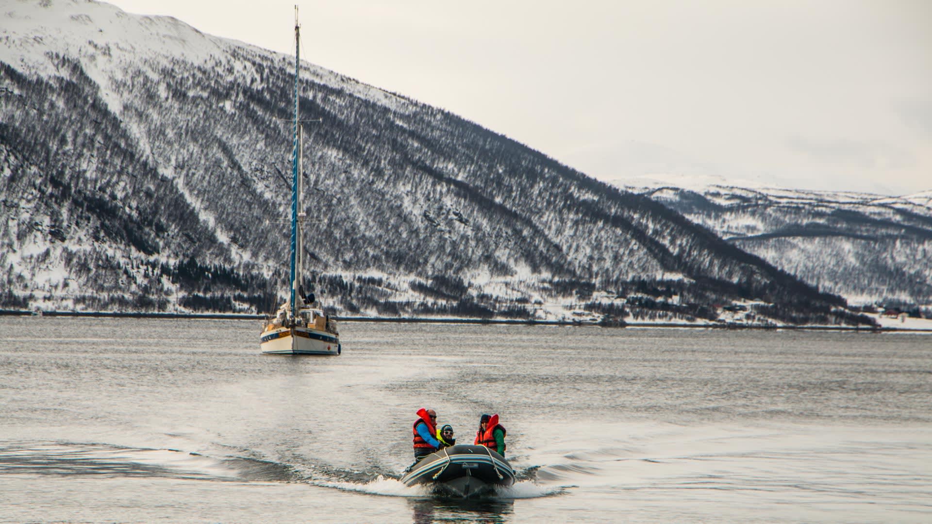 Drei Skitourengeher im Schlauchboot fahren auf einem Fjord in Norwegen zu einem Segelboot.