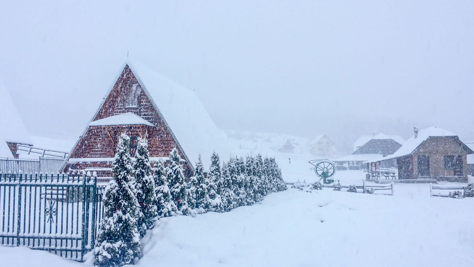 Schneebedeckte Holzhäuser und Nadelbäume bei starkem Schneefall in einer Wintersiedlung in Montenegro.