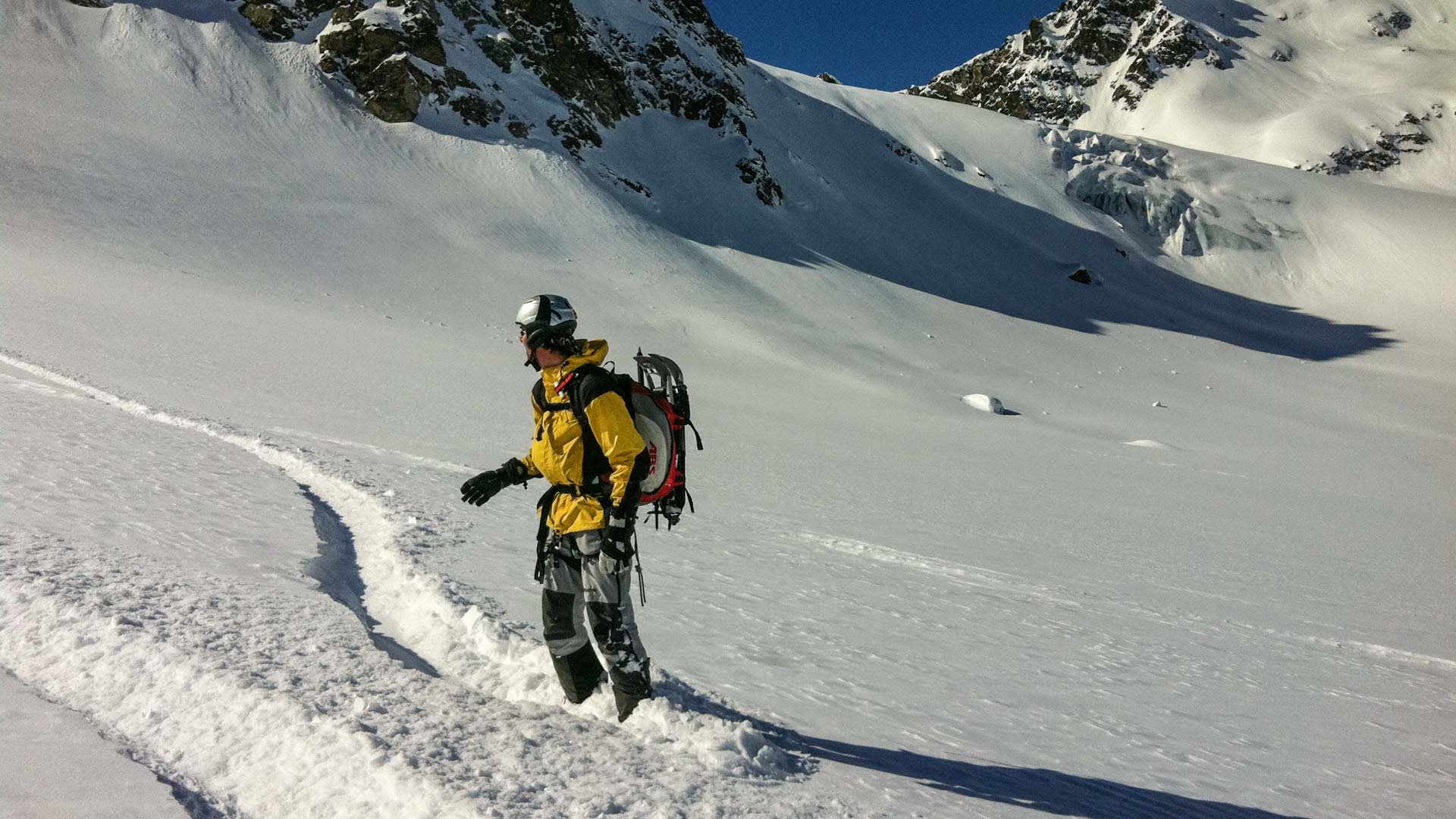 Person mit Splitboard und Airbag-Rucksack steht in Tiefschneespur vor gletscherbedecktem Steilhang bei Skitour am Gletscher.