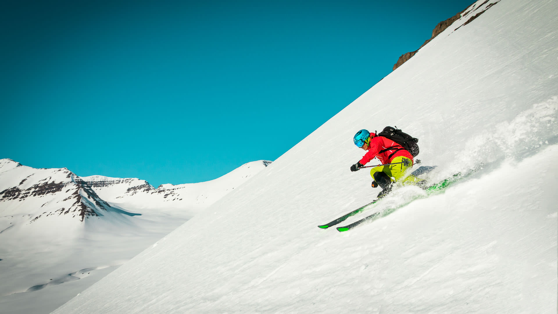 Skitourengeher in roter Jacke fährt stark geneigten Schneehang auf Island mit aufgewirbeltem Schnee ab.