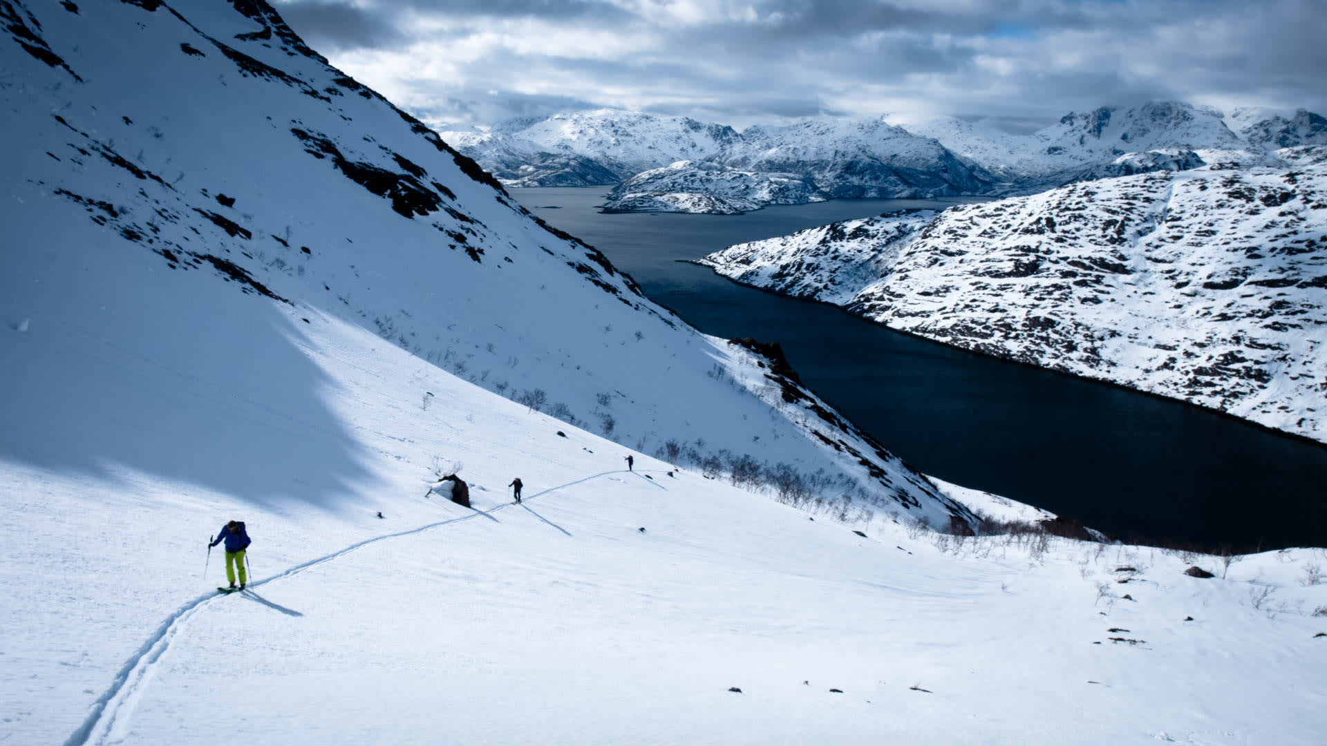 Vier Skitourengeher steigen in einer Spur einen verschneiten Steilhang in Nordnorwegen auf, im Hintergrund ein Fjord.