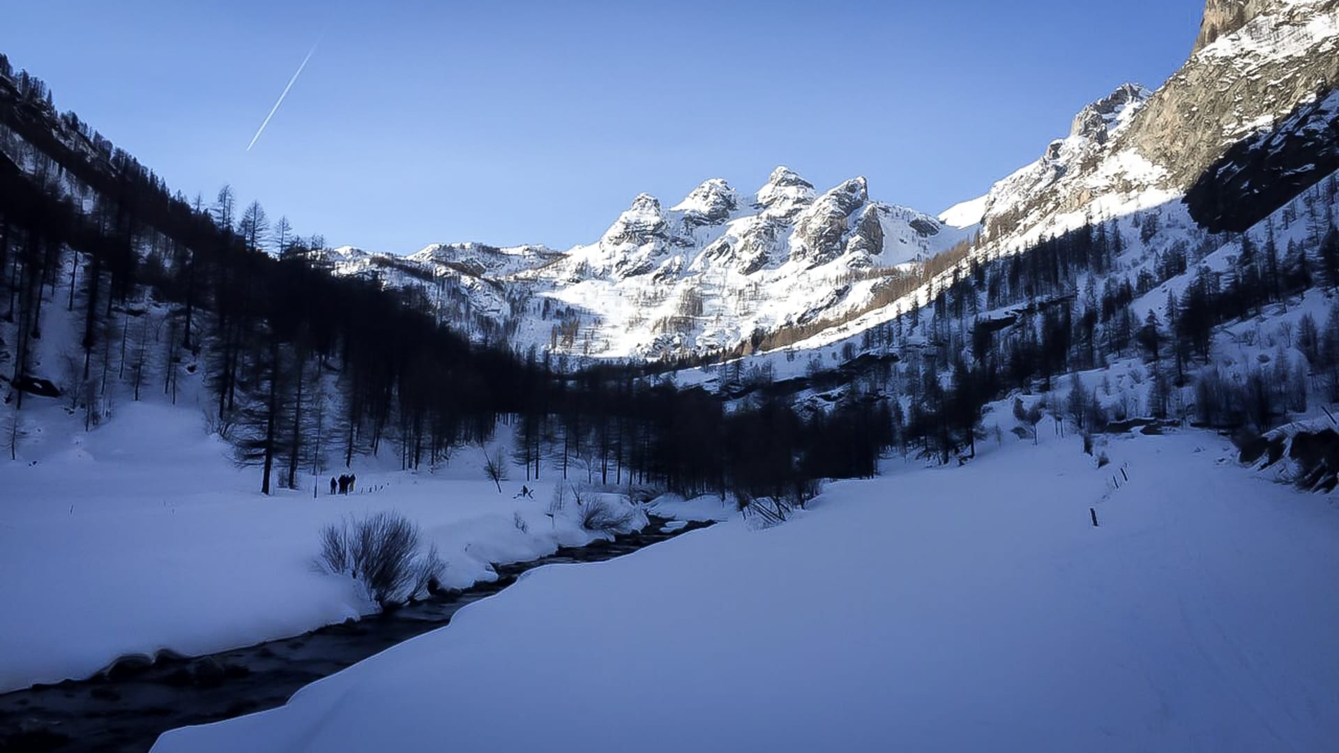 Drei Skitourengeher in der Talsohle des Valle Maira vor einer schneebedeckten Felsgipfelgruppe in Piemont.