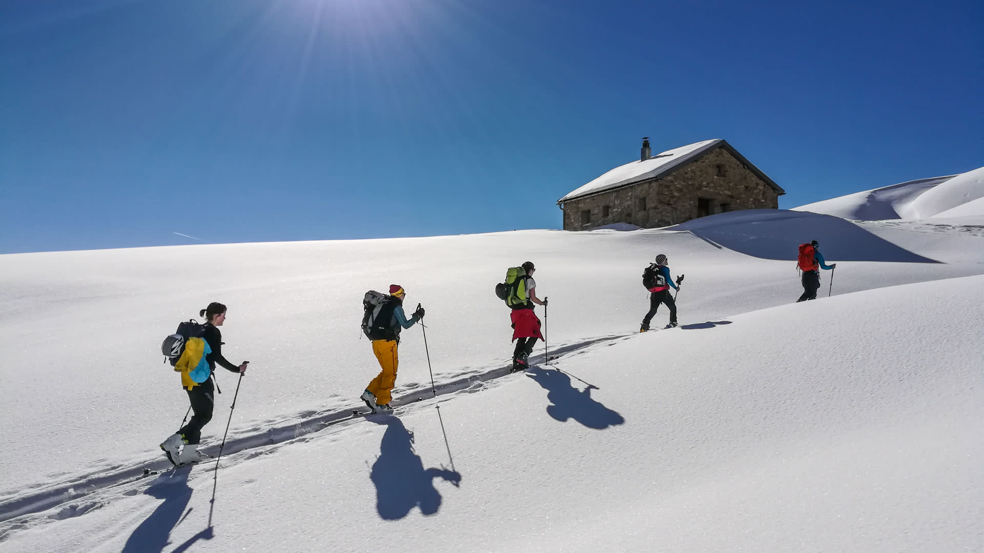 Fünf Skitourengeher steigen mit Fellen auf einem verschneiten Hang im Vinschgau auf, im Hintergrund eine Steinalpenhütte