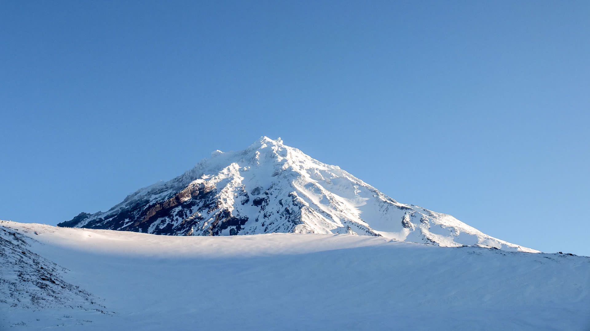 Kegelförmiger, schneebedeckter Vulkangipfel in Kamtschatka mit weitem Schneefeld im Vordergrund und klarem Winterhimmel.