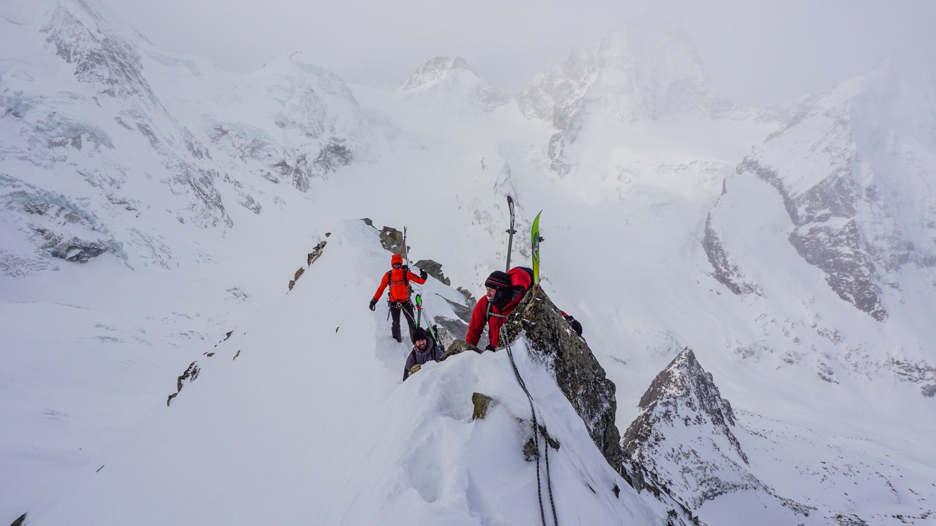 Drei Personen mit Ski am Rücken klettern an einem Fixseil über einen schmalen Schneegrat bei der Skitour im Wallis.