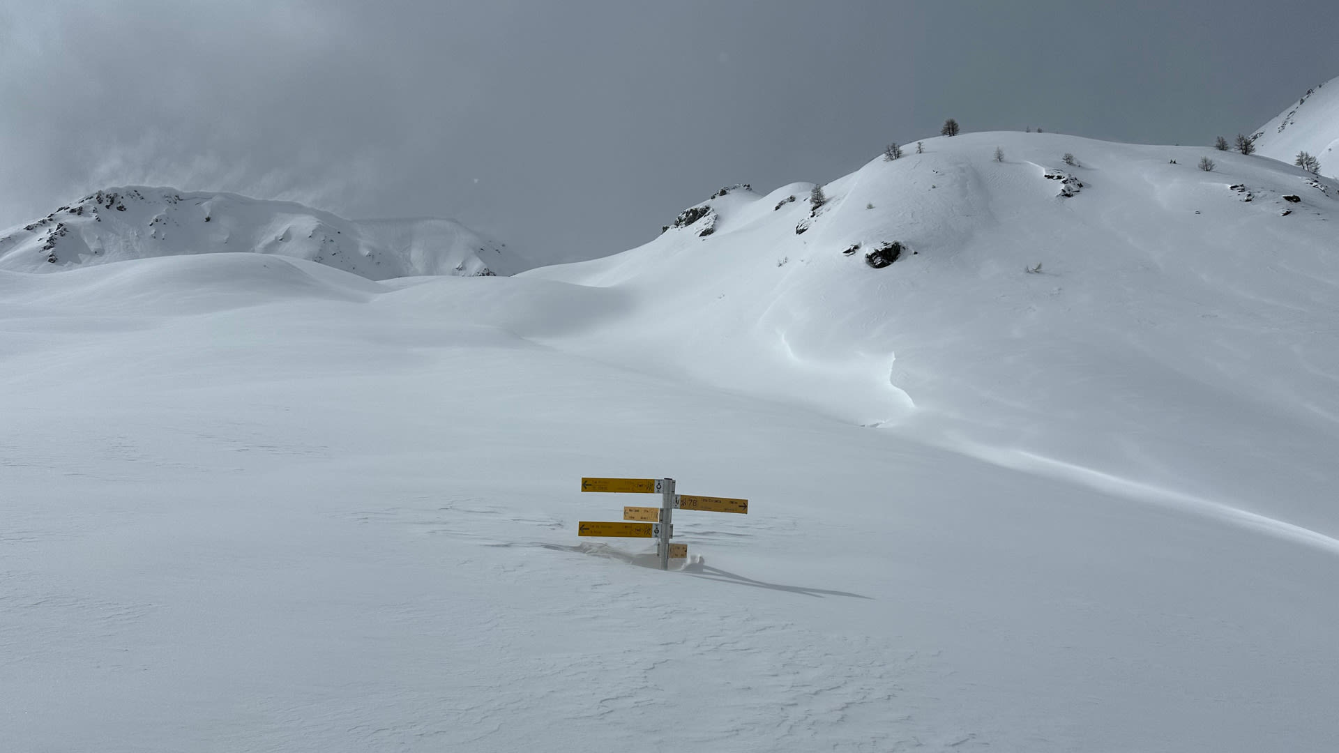 Halb eingeschneiter gelber Wegweisermast auf einer Skitour in den Walliser Alpen bei bedecktem Winterhimmel.