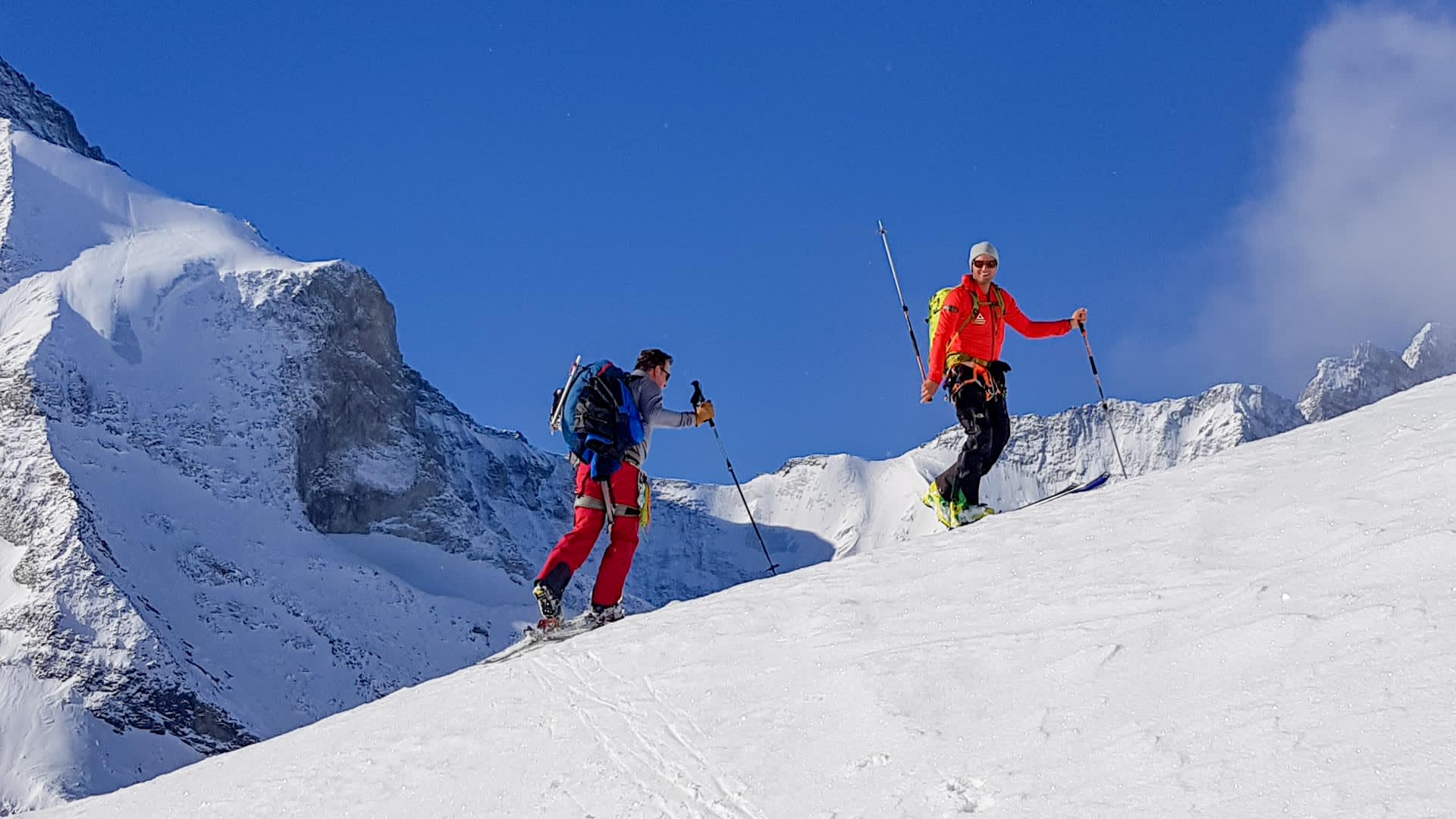 Zwei Skitourengeher steigen mit Fellen einen steilen Schneehang im Wallis auf, im Hintergrund ein markanter Felsturm.