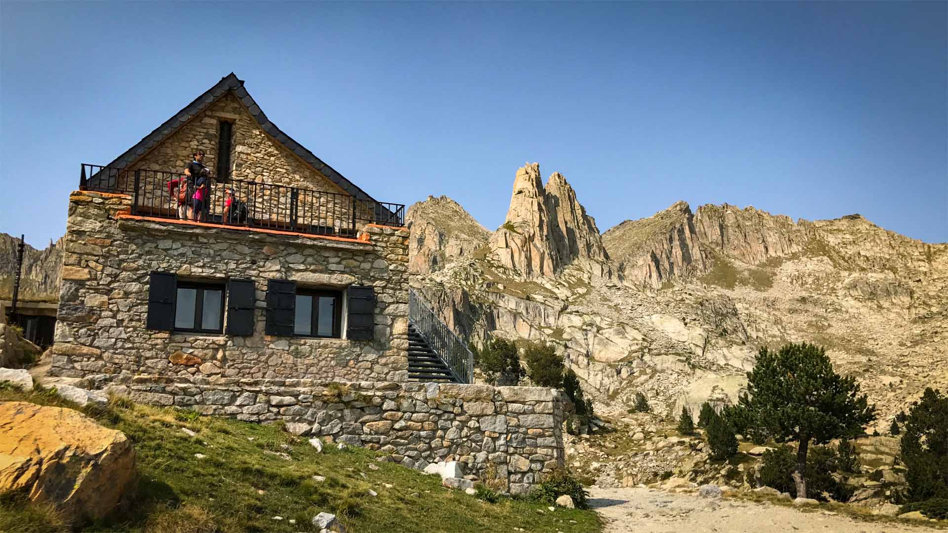 Trekking am Refugi d'Amitges bei Espot: Natursteinschutzhaus mit Trekkern auf dem Balkon, dahinter zerklüftete Felstürme ohne