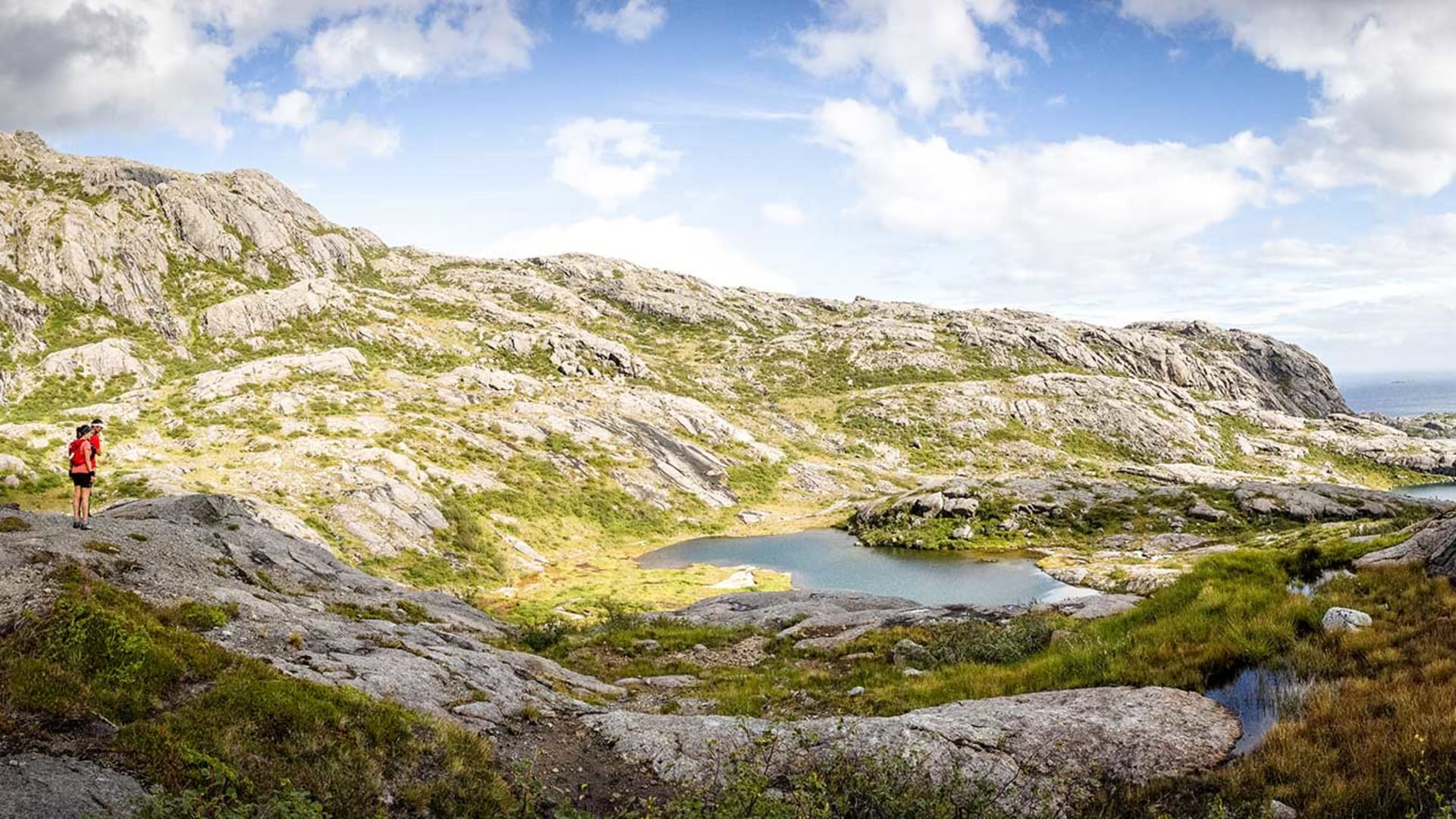 Trekking auf den Lofoten: zwei Trekker mit roten Jacken auf Granitplateau mit Blick auf kleinen Bergsee und Fjord.