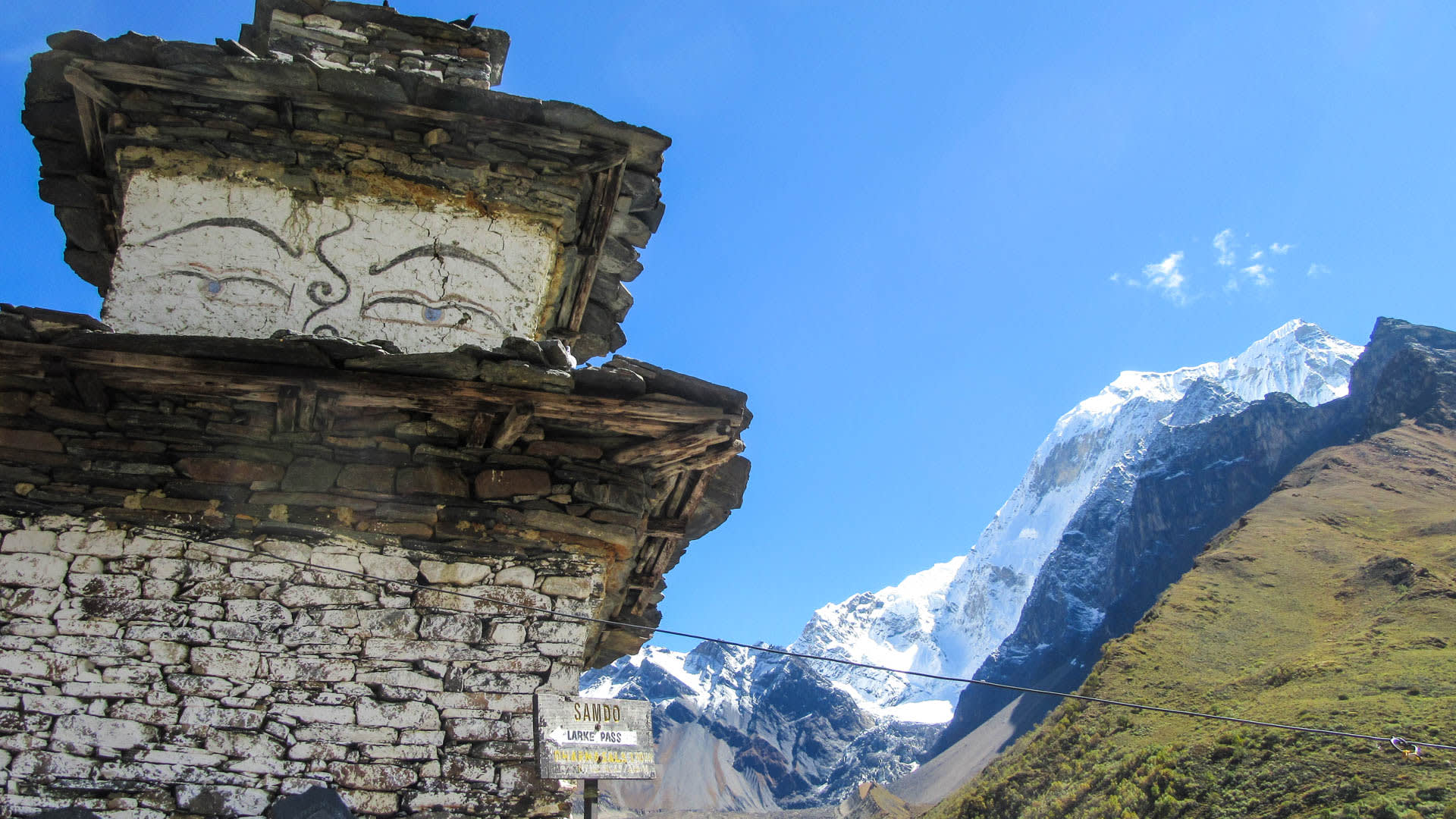 Trekking am Manaslu Trek: verwitterter Chorten bei Samdo mit Buddha-Augen und Schild zum Larkya Pass vor vergletschertem Gipfel.