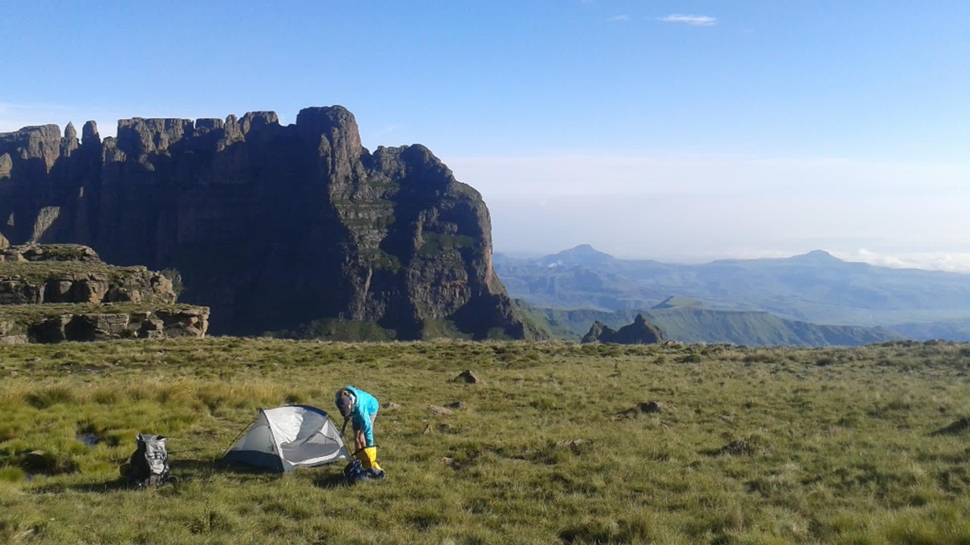 Trekker baut Kuppelzelt auf dem Hochplateau der Drakensberge auf, großer Rucksack liegt daneben, steile Felswand im Hintergrund.