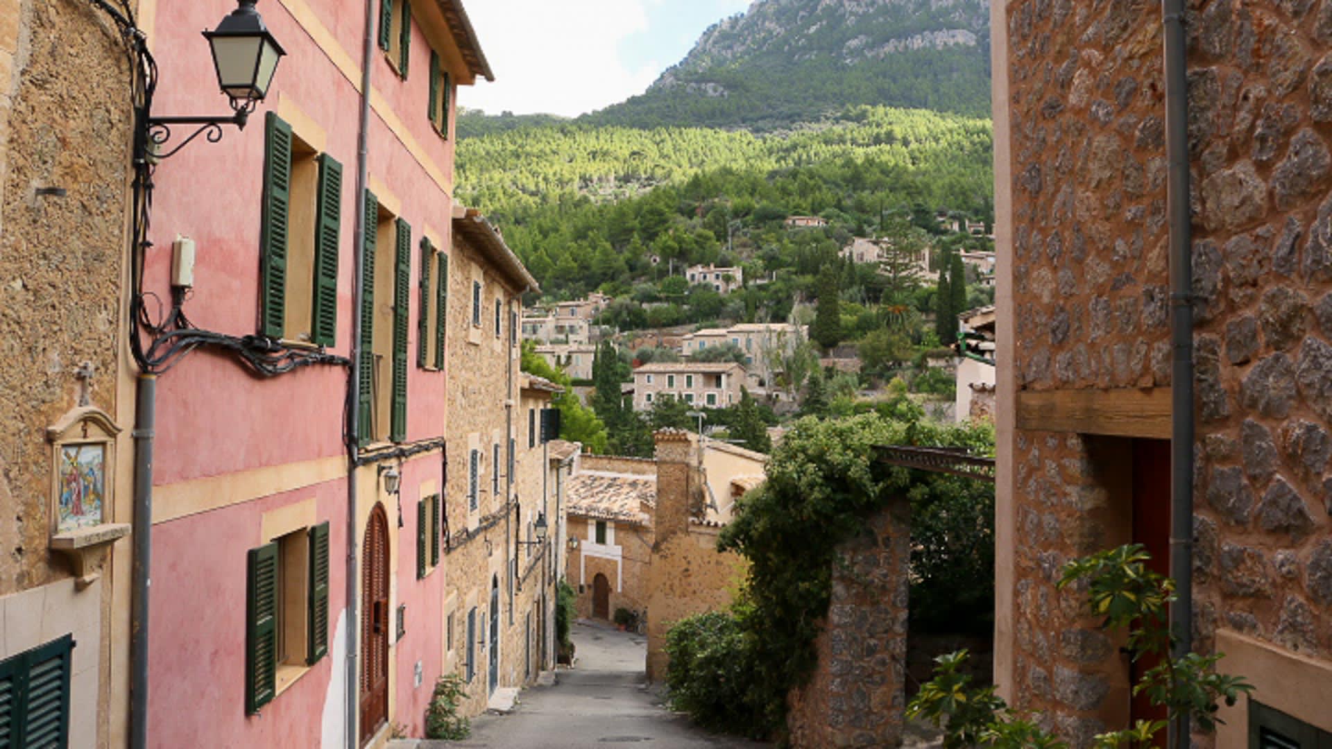 Trekking in Sóller: gepflasterte Gasse mit Natursteingebäuden, grünen Fensterläden und Blick auf den felsigen Gebirgskamm dahinter.