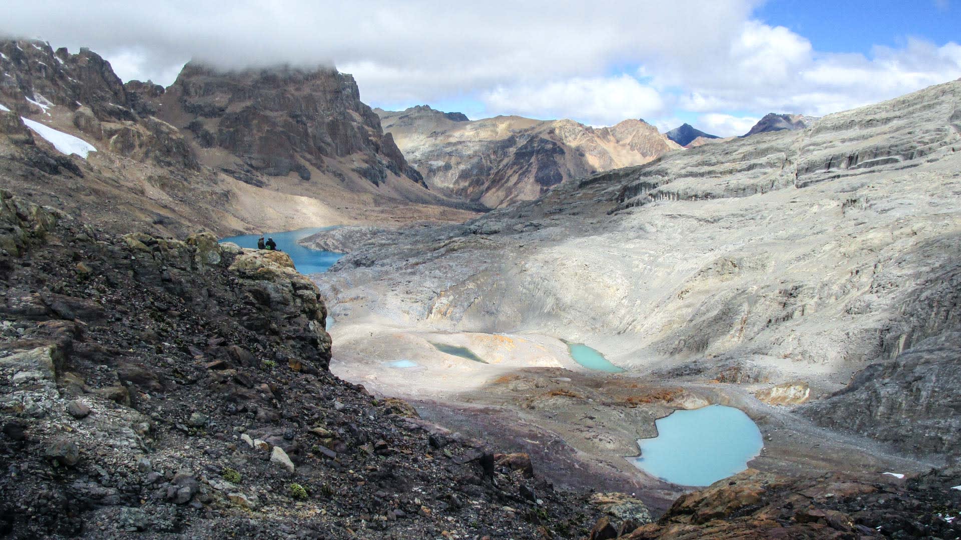 Zwei Trekker rasten auf Felsvorsprung über glazialem Becken mit türkisen Gletscherseen zwischen Carhuacocha und Huayhuash.