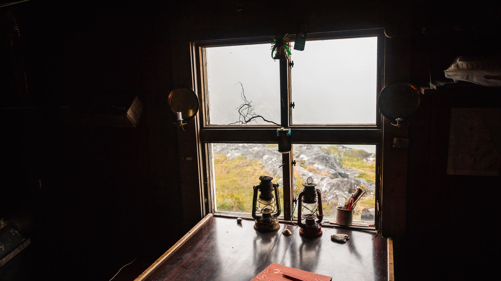 Innenraum einer rustikalen norwegischen Hytta mit Petroleumlampen, Holztisch und Blick durch ein Fenster auf Felslandschaft.
